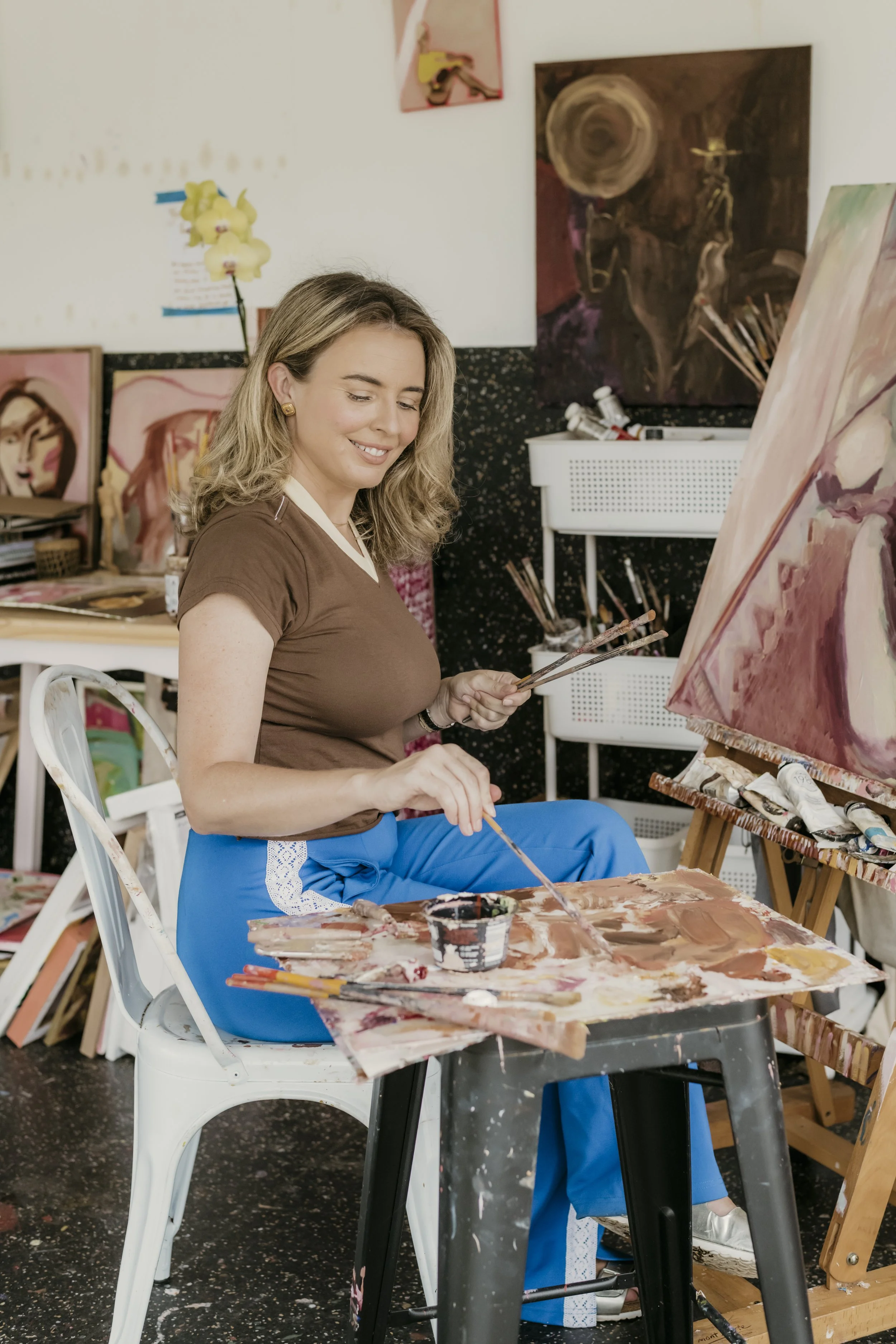 A woman in a brown shirt and blue pants sitting on a white chair, painting on a canvas in an art studio. The studio has paintings on the wall, art supplies, and a small table with paint and brushes.