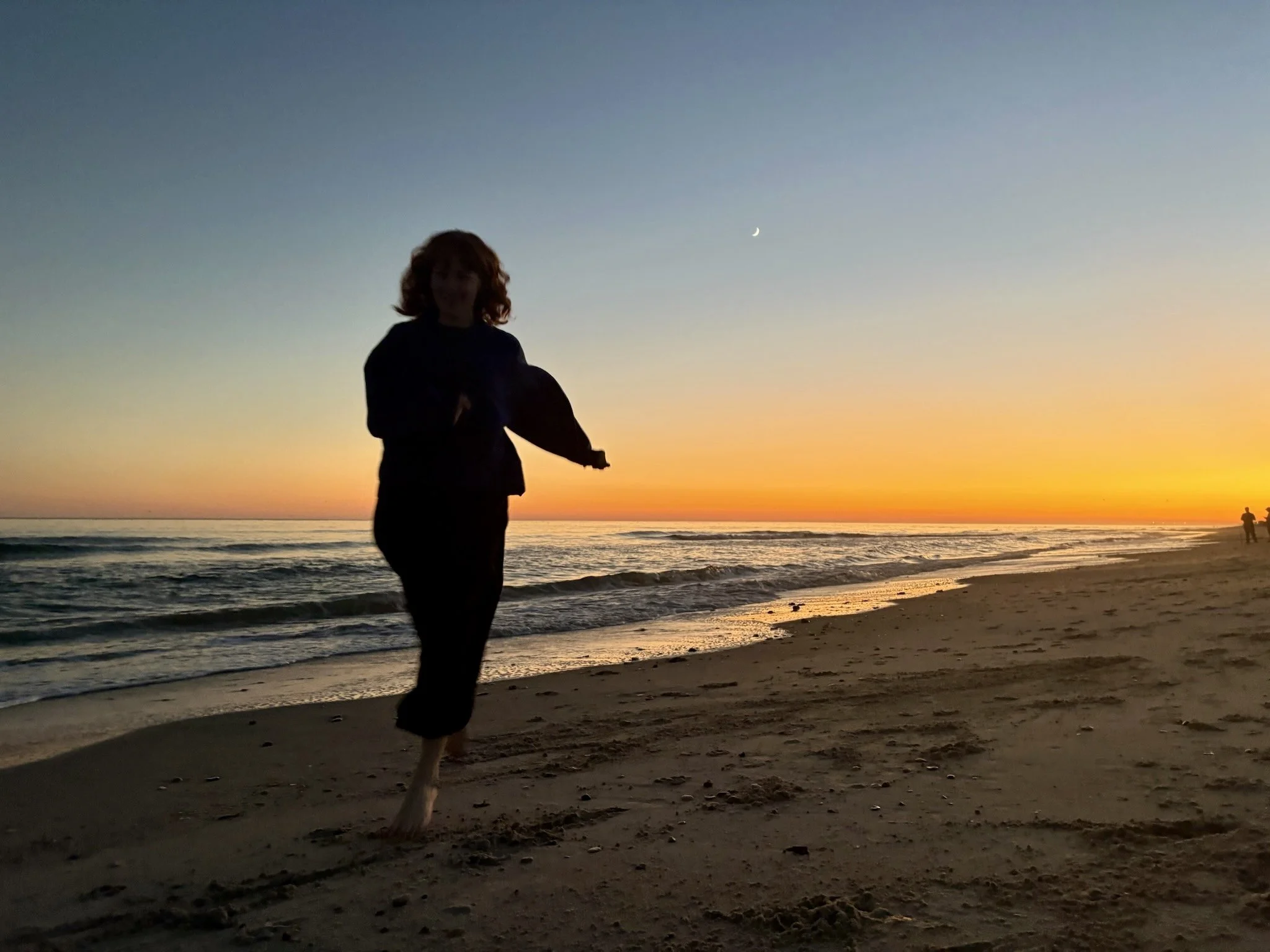 A person with red hair walking on the beach during sunset with a small crescent moon in the sky and another person further away by the water.