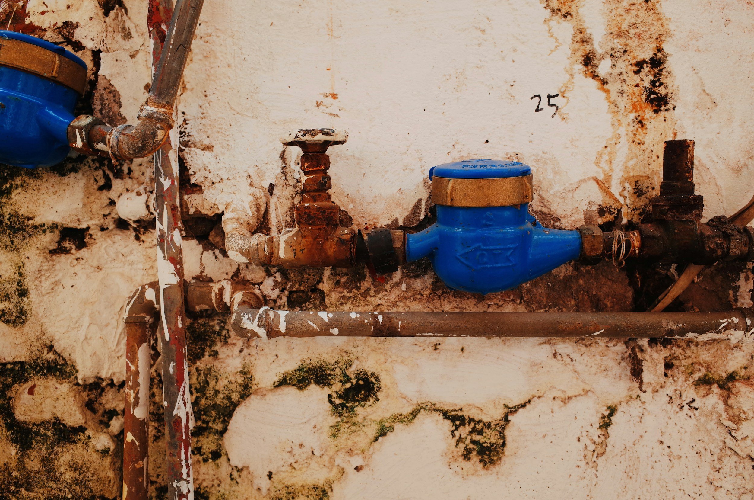 Rusty plumbing pipes and water meters attached to a weathered, stained wall with peeling paint.