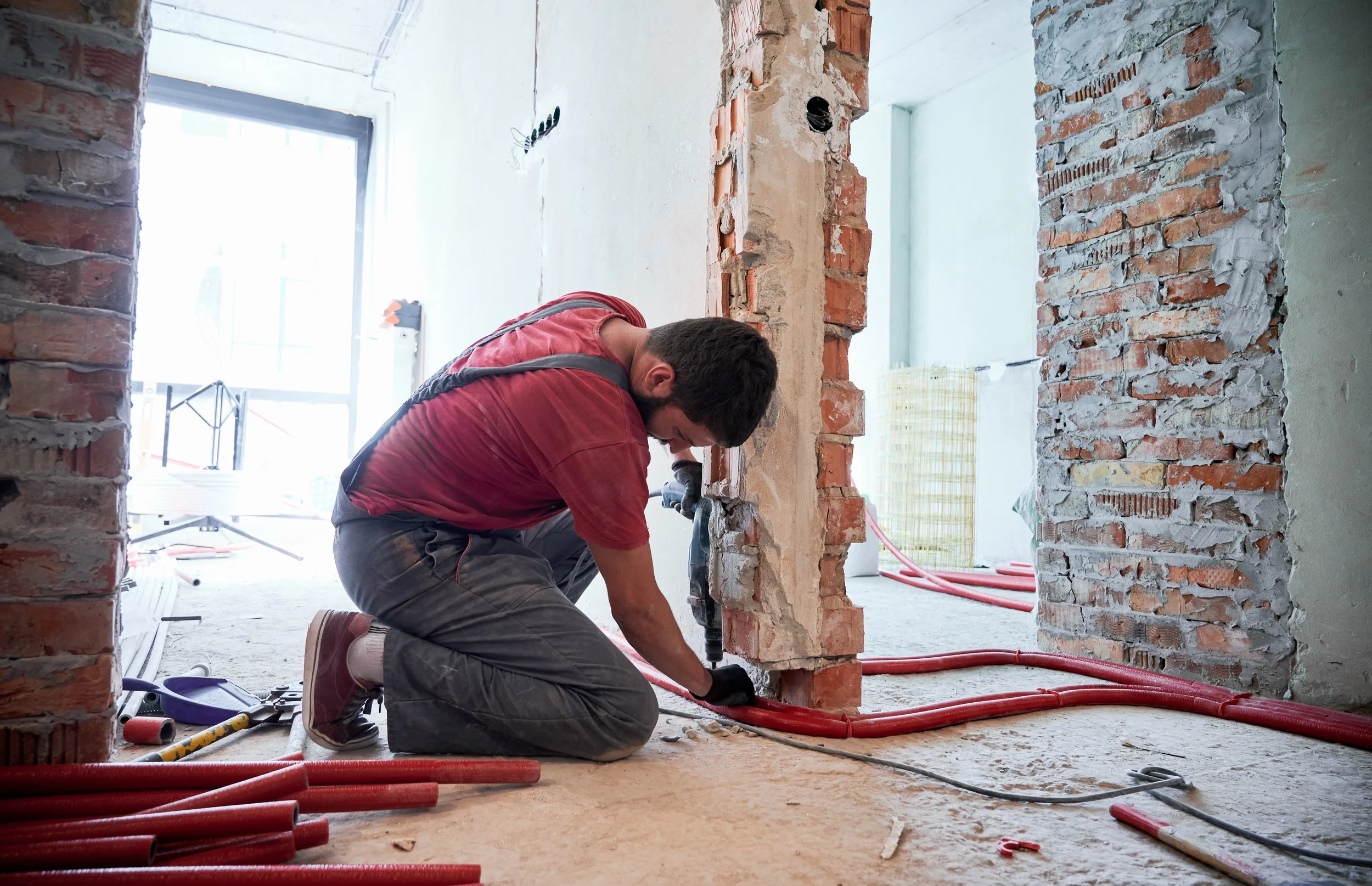 A man working on construction, kneeling on the floor and using a power drill on a brick wall inside a building under renovation.