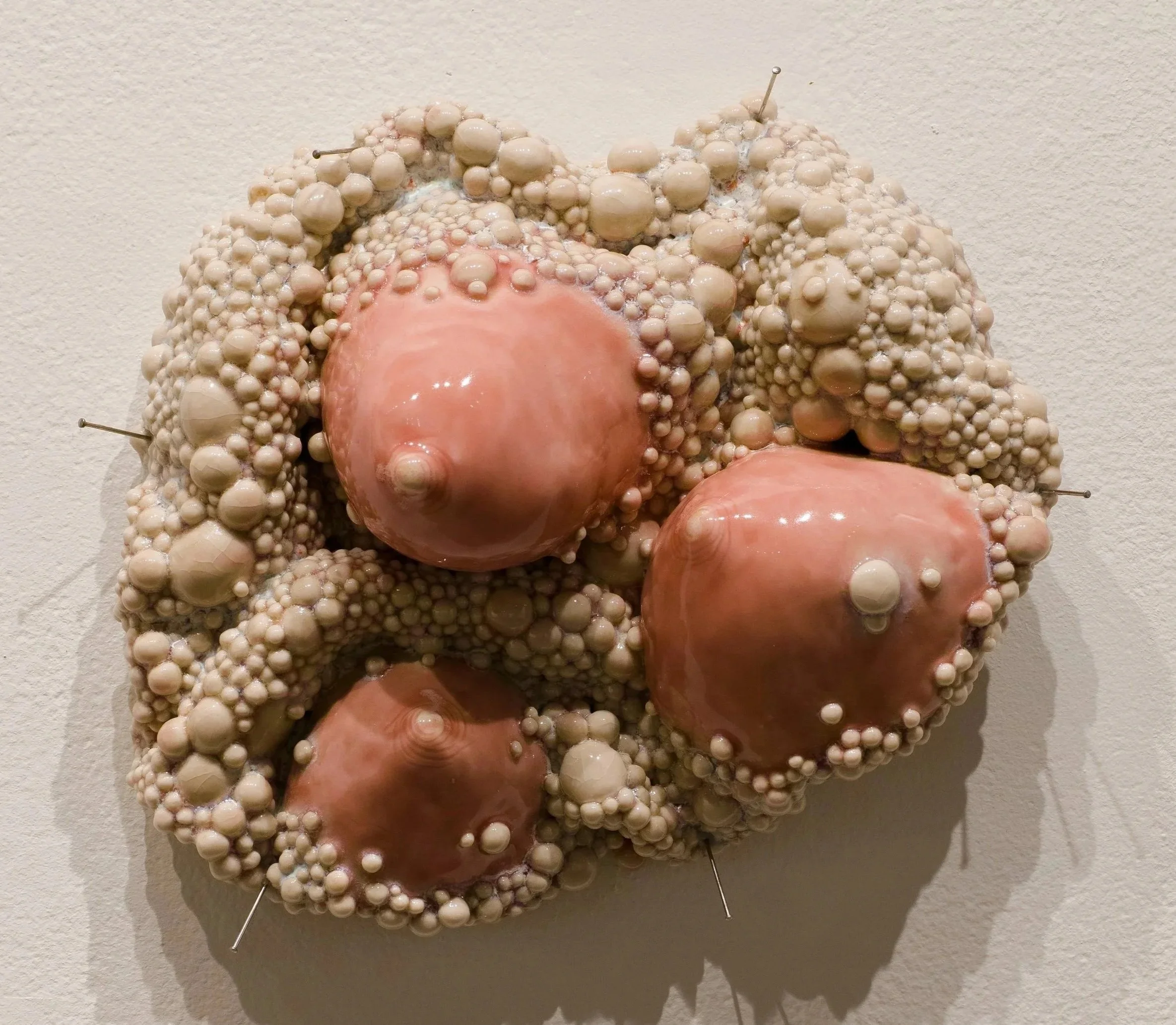 Ceramic wall sculpture of breasts surrounded by cream bumpy diseased flesh.