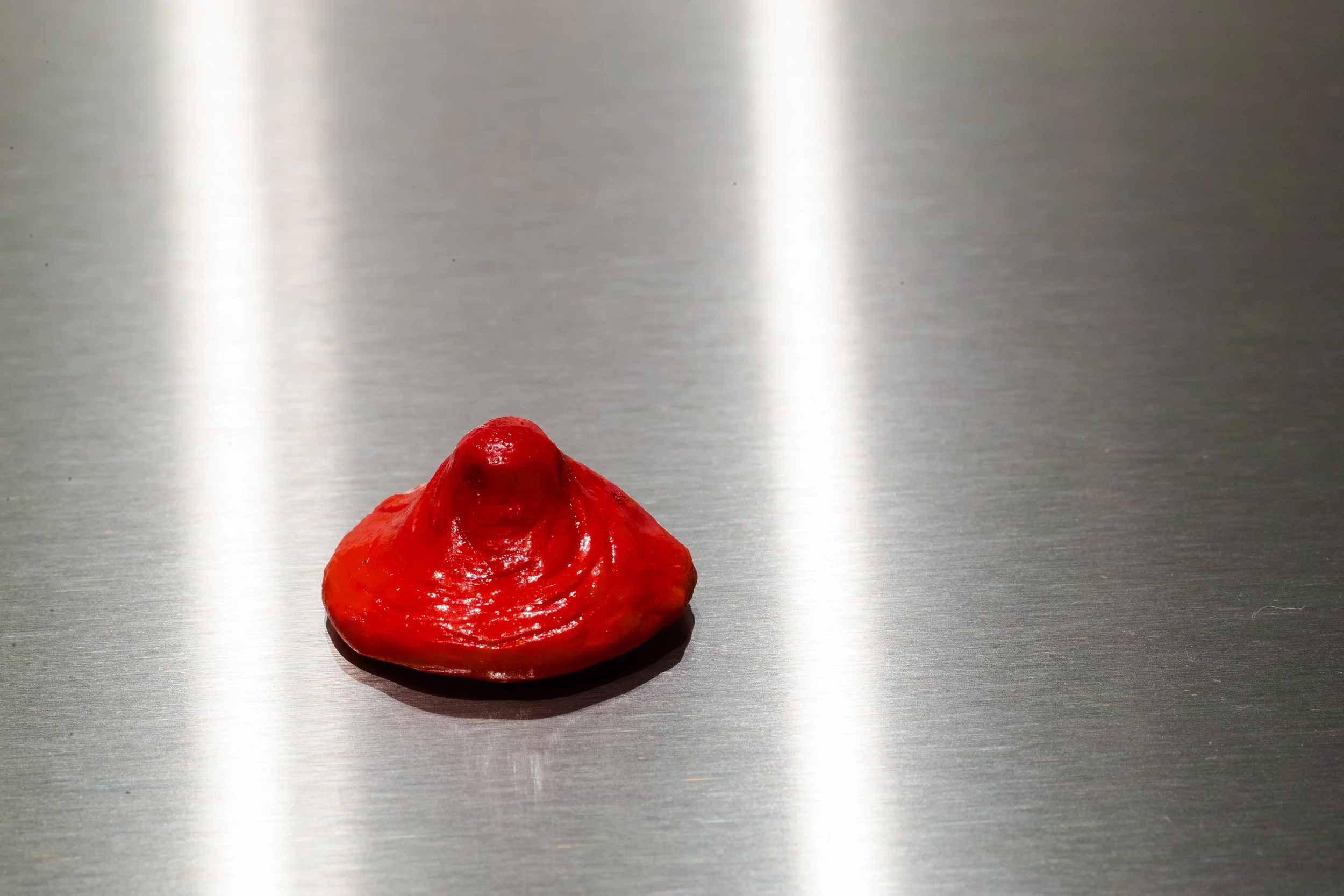 A red substance with a glossy texture, shaped like a small mountain or peak, placed on a reflective metallic surface.