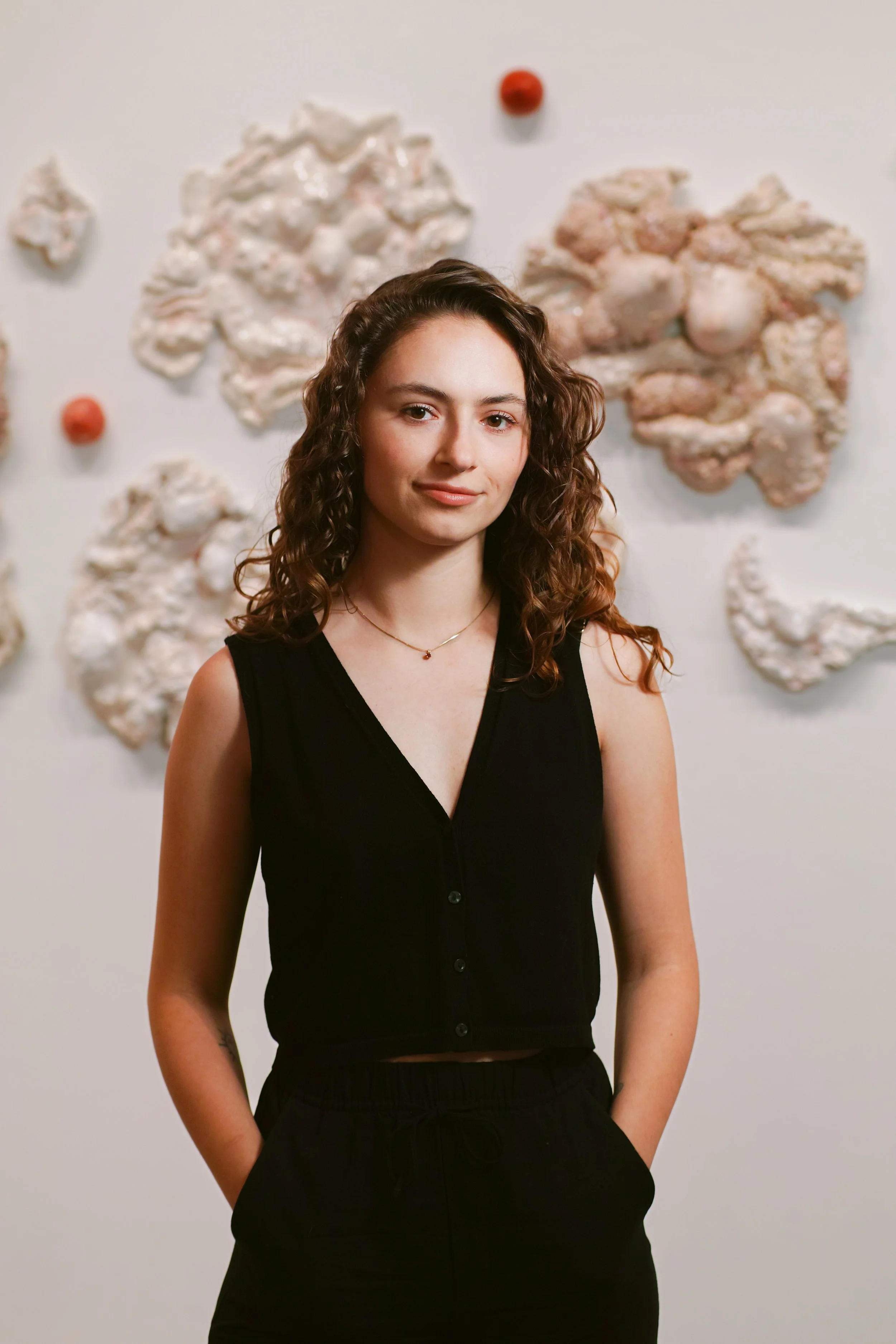 A young woman with long, curly brown hair, wearing a black sleeveless button-up top, standing in front of a white wall decorated with abstract, textured, cream-colored shapes and small orange spherical objects.