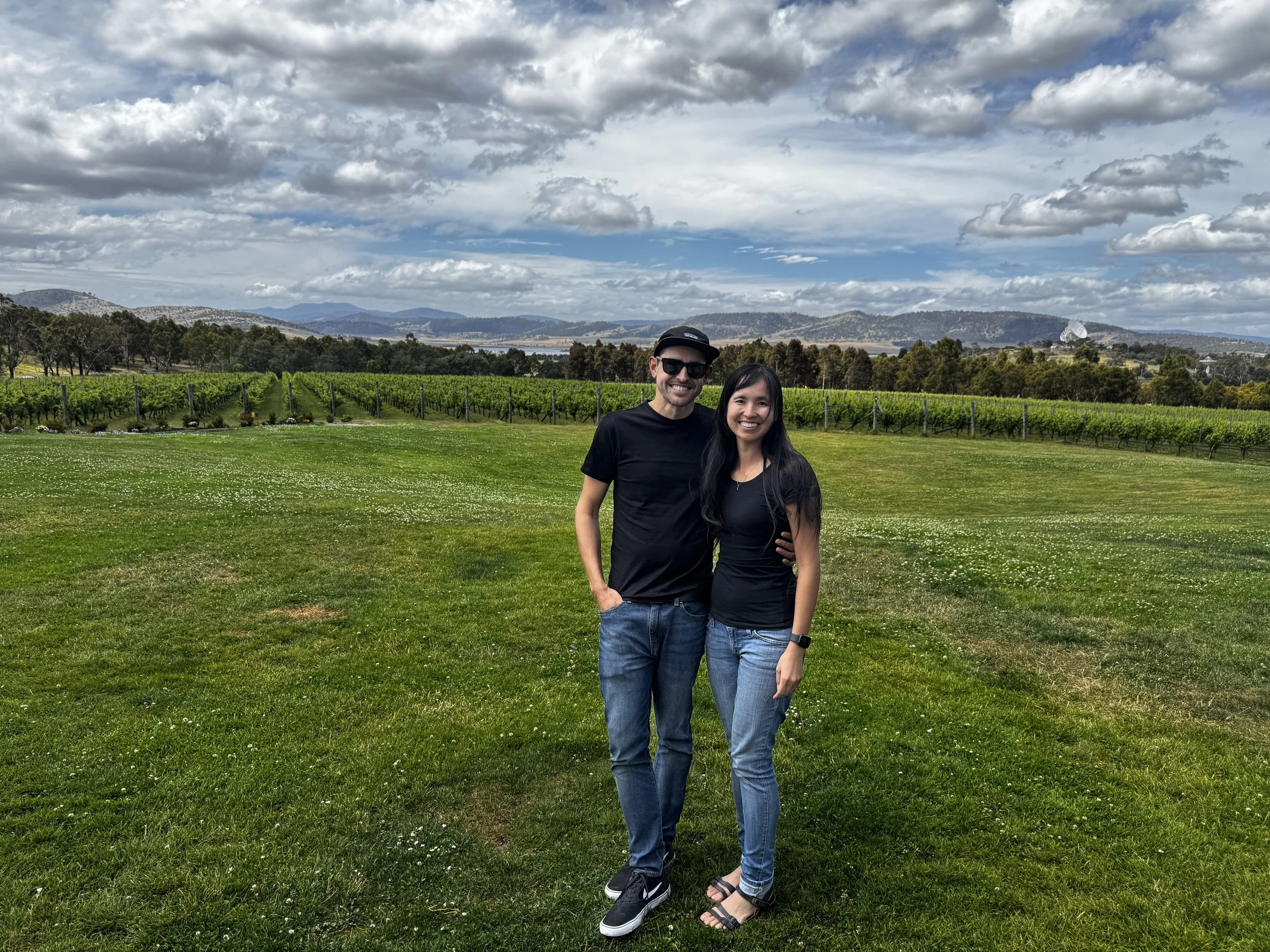 A smiling man wearing sunglasses, a cap, and a black t-shirt stands beside a smiling woman with long dark hair, wearing a black top and jeans, in a scenic vineyard with rolling hills and a partly cloudy sky in the background.