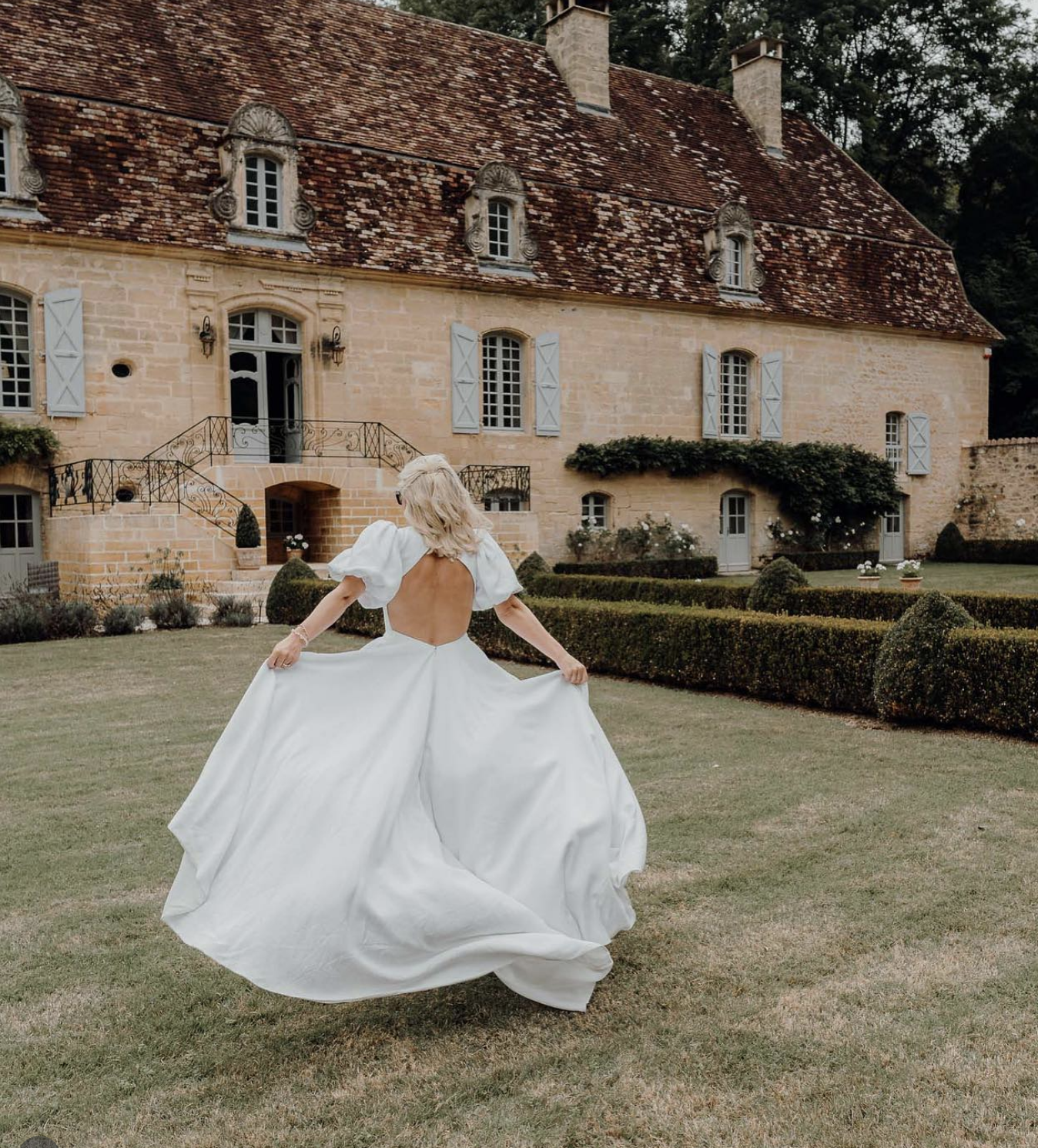 Woman in a white wedding dress dancing or twirling on a green lawn in front of a large, old stone house with a steep, red-tiled roof and multiple windows with light blue shutters.