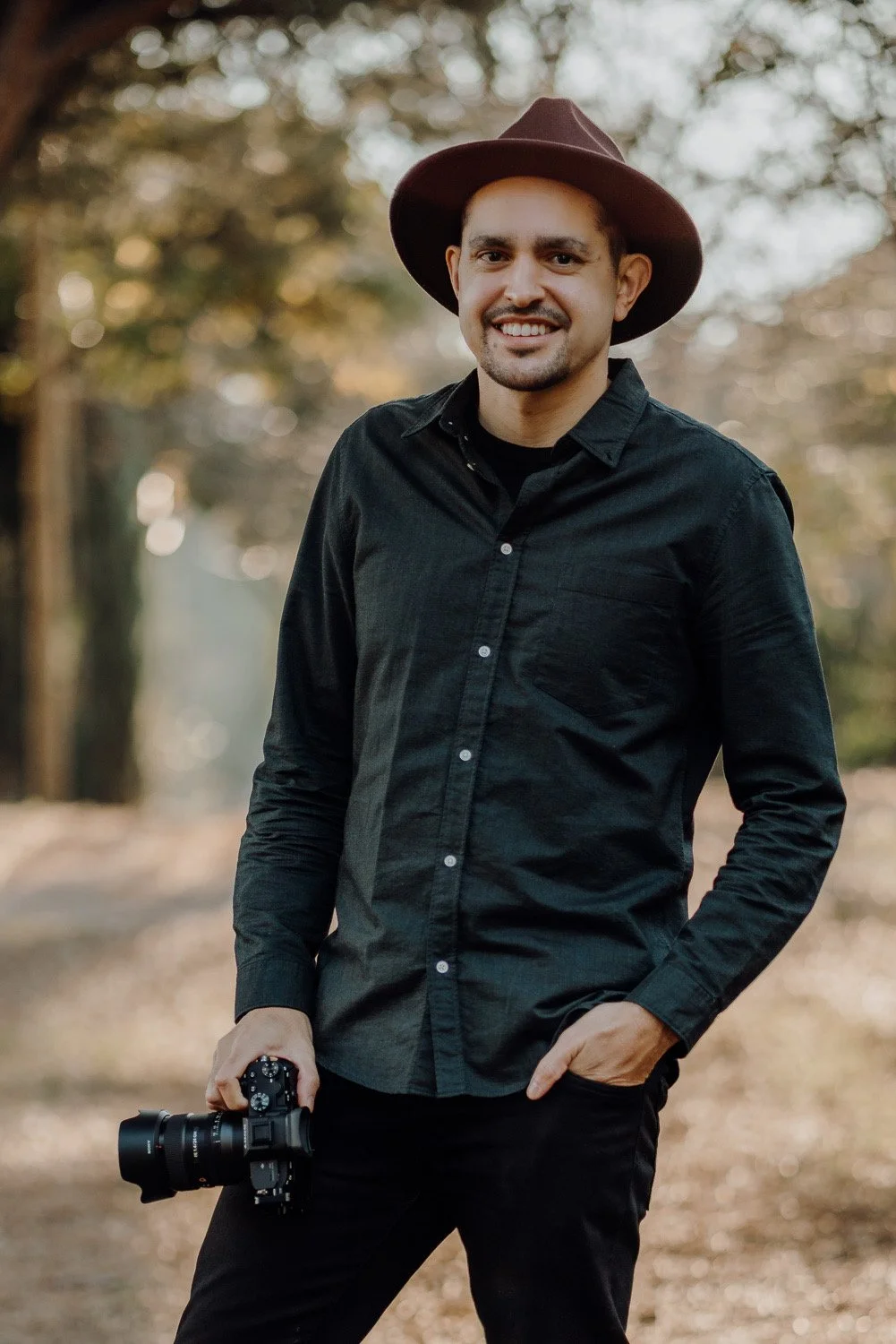 A man smiling outdoors wearing a black shirt and a brown wide-brimmed hat, holding a professional camera.