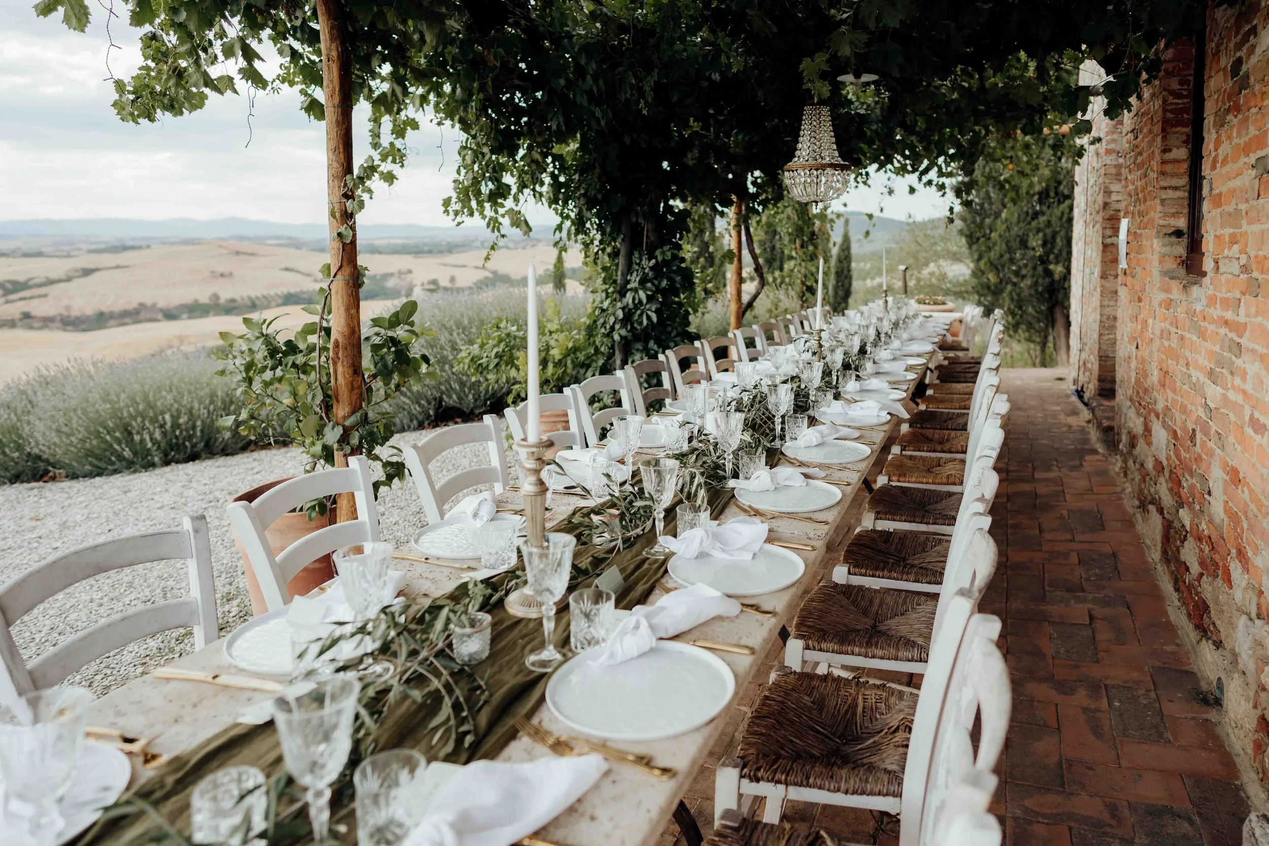 An outdoor dining setup with a long table covered with a white floral tablecloth, set with plates, glasses, and silverware. The table is decorated with white and yellow flowers. The dining area is along a dirt path with string lights hanging overhead, and is surrounded by green plants and climbing vines. In the background, there is a hill with a rustic wooden building and a tall tree, with soft sunlight illuminating the scene.