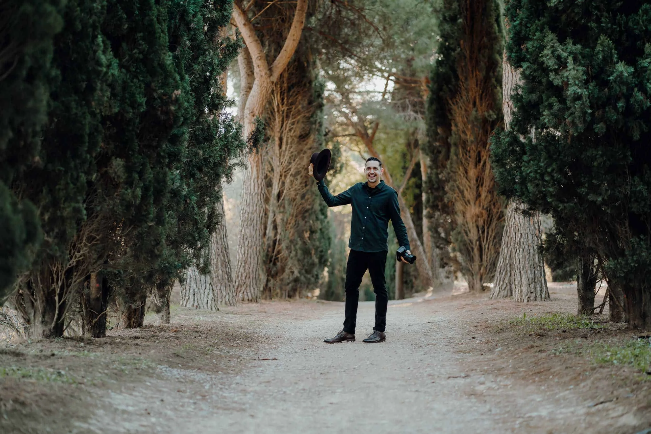 A man wearing a black long sleeve shirt and dark pants standing on a dirt path between tall trees, holding a black hat in his right hand and a camera in his left hand, smiling and looking towards the camera.
