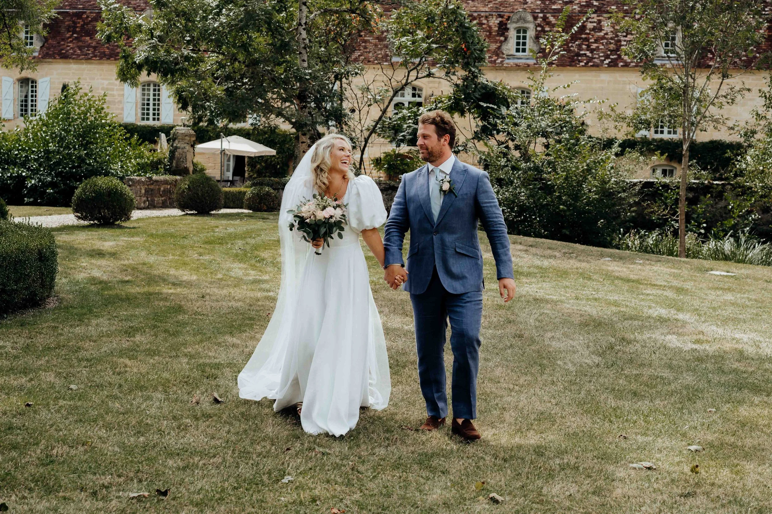 A bride and groom holding hands and walking together on a lawn during their wedding, with a large historic house and lush green trees in the background.