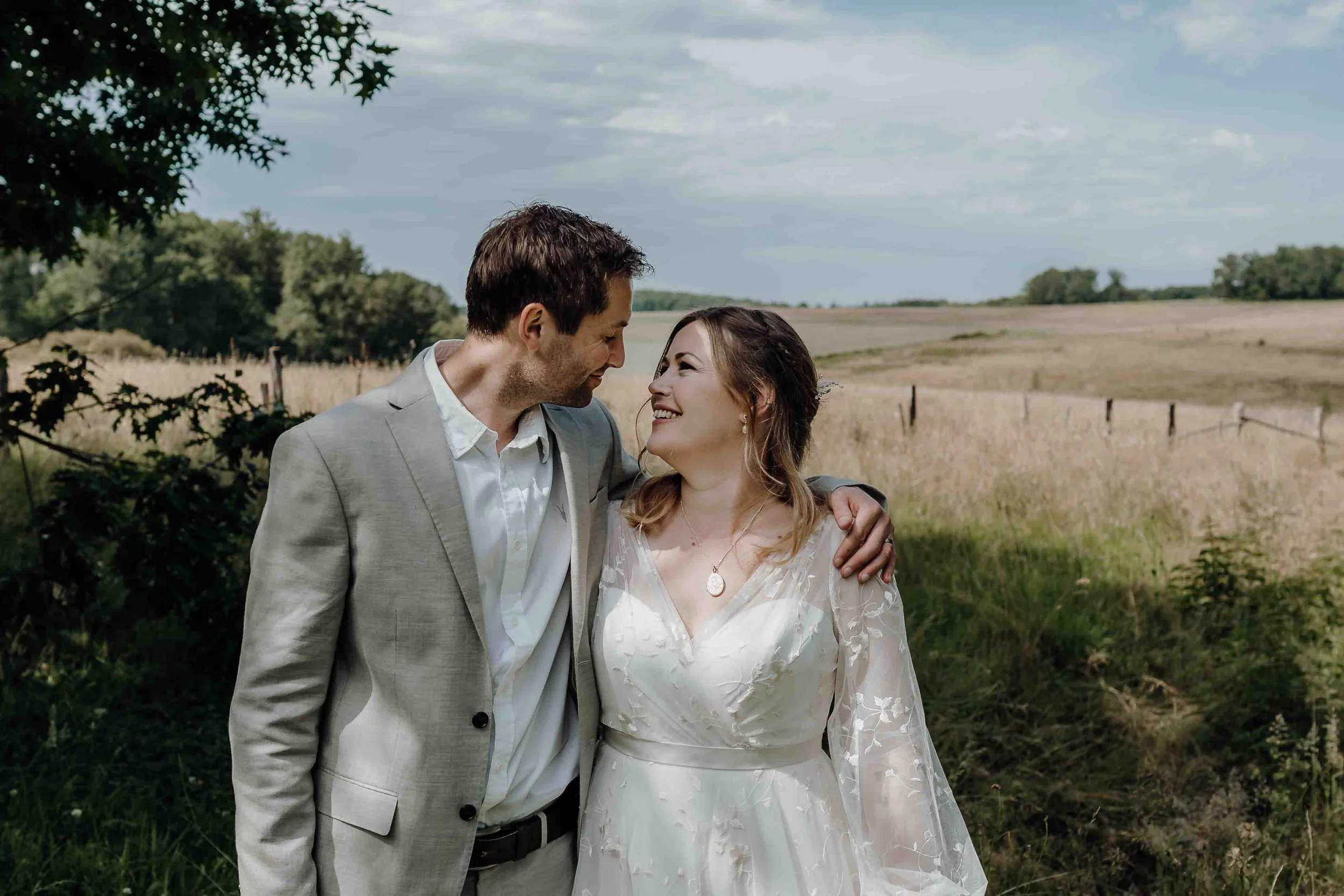 A bride and groom standing together outdoors on a grassy area near a field, with tall green trees in the background, smiling and facing each other.
