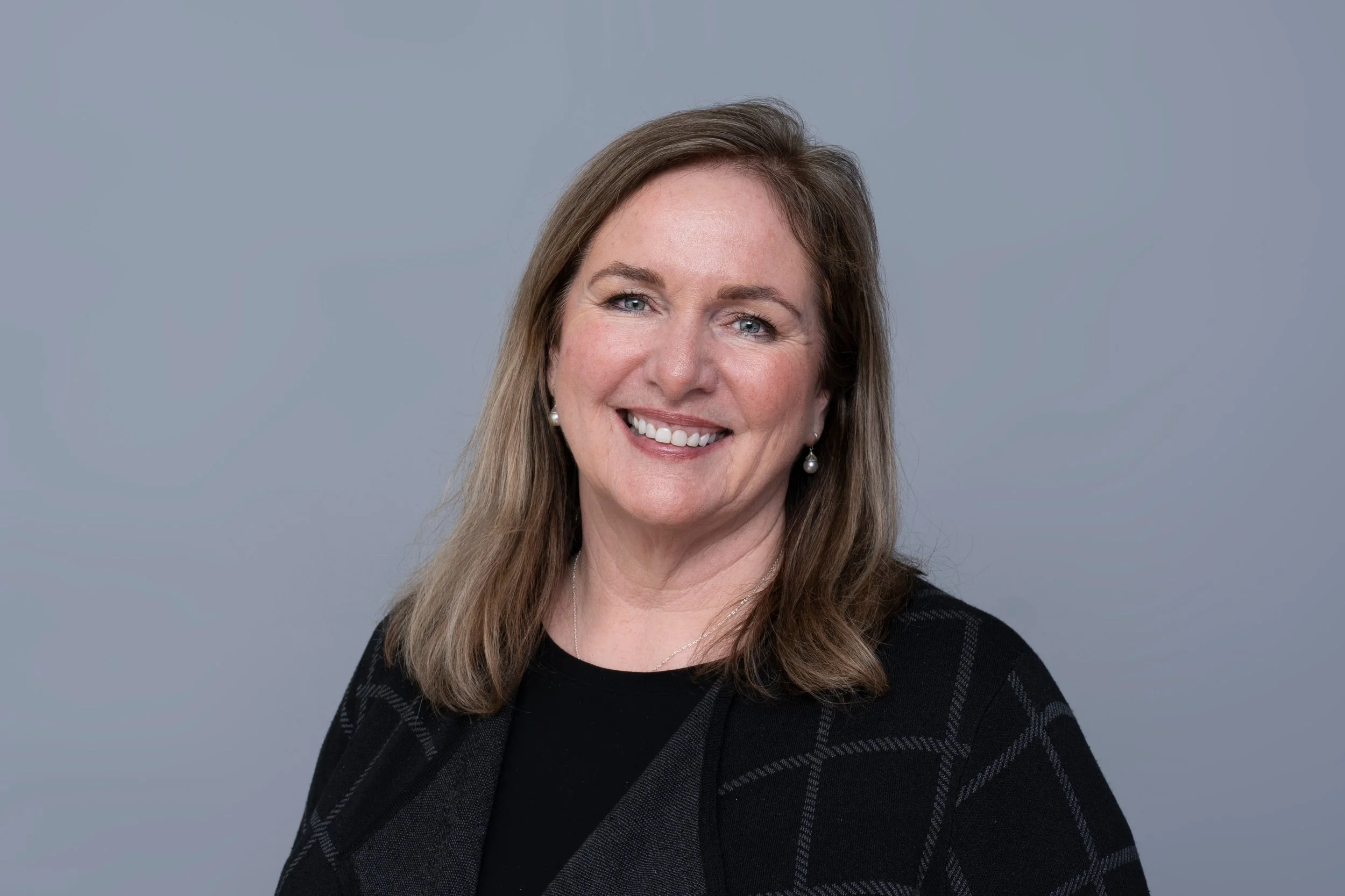 A woman with long light brown hair, blue eyes, and pearl earrings smiling against a plain gray background.