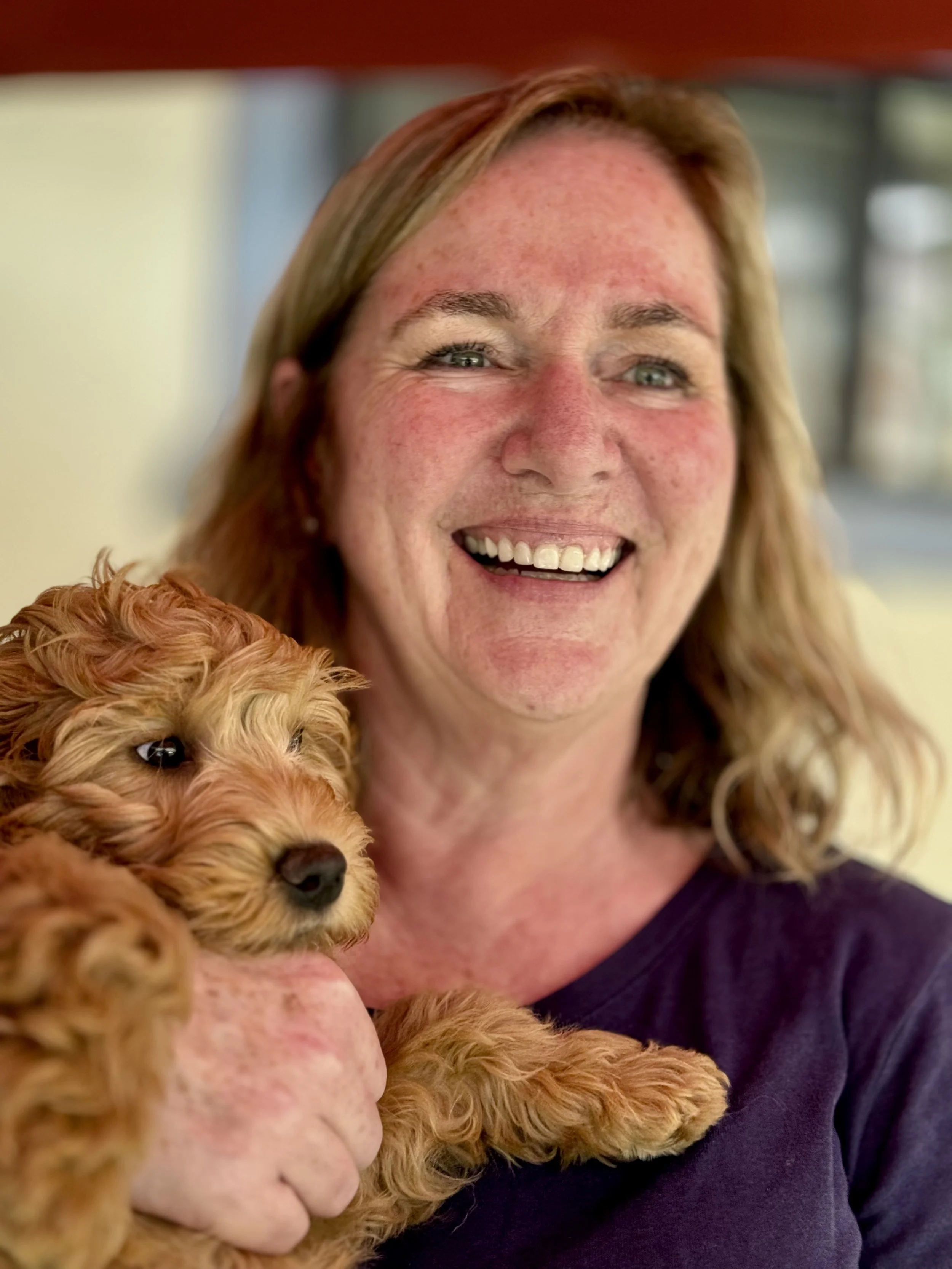 A woman with red hair and a dog with curly fur are smiling and posing together for a photo indoors.