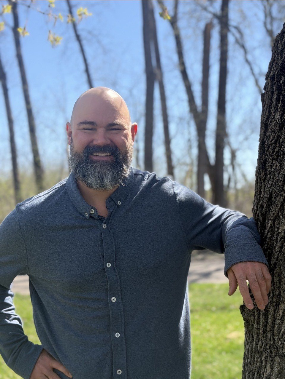 A smiling man with a beard wearing a dark button-up shirt standing outdoors near a tree in a park during daytime with trees and a blue sky in the background.