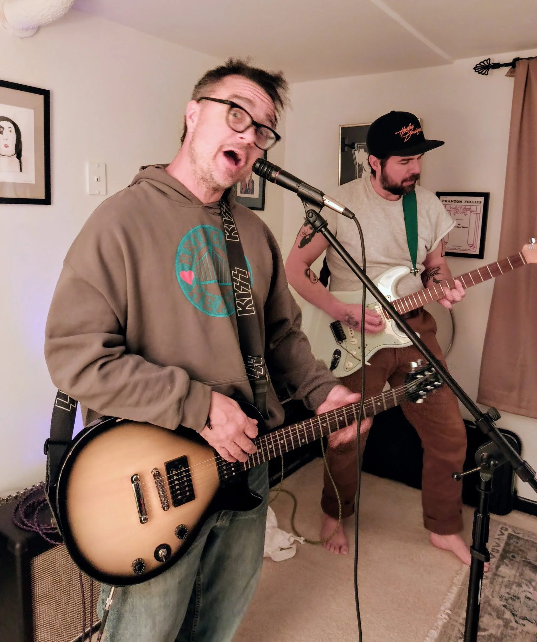 Two men playing musical instruments in a cozy indoor setting. The man in the foreground wears glasses and a hoodie, singing into a microphone while holding an electric guitar. The man in the background, with a beard and wearing a cap, is focused on playing another guitar. The room is decorated with framed artwork on the walls, a patterned rug, and a beige curtain.