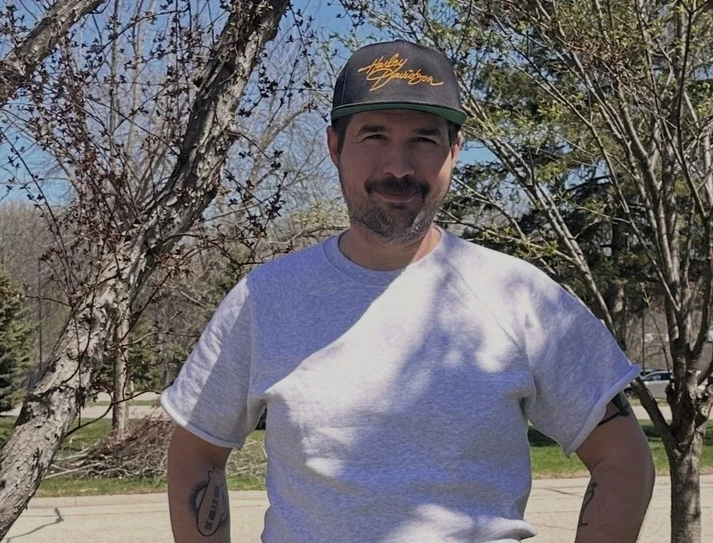 Tim Liddicoat standing outdoors on a sidewalk, wearing a gray t-shirt and a black cap with gold lettering, surrounded by trees with some leaves, on a sunny day.