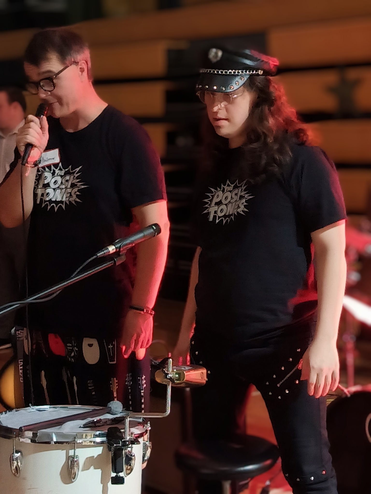 Young woman playing drums at a music event, wearing glasses, a black t-shirt with a comic-style text design, and a police cap, standing next to a drum set.