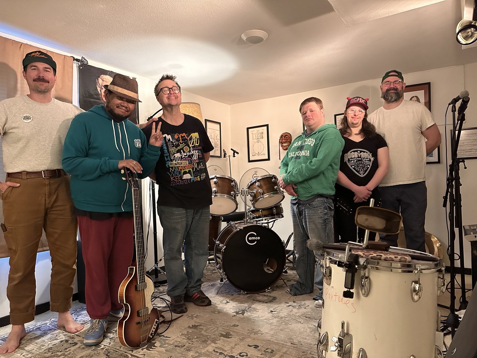 Group of seven people in a room with musical instruments, including drums, guitar, and microphone, posing for a photo.