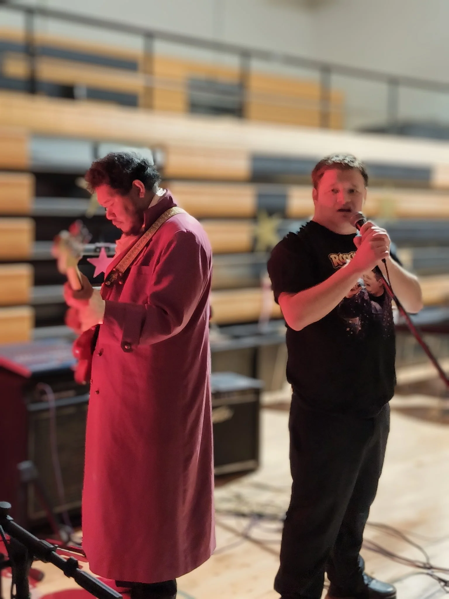 Two men performing with microphones in a room with stacked bleachers in the background.