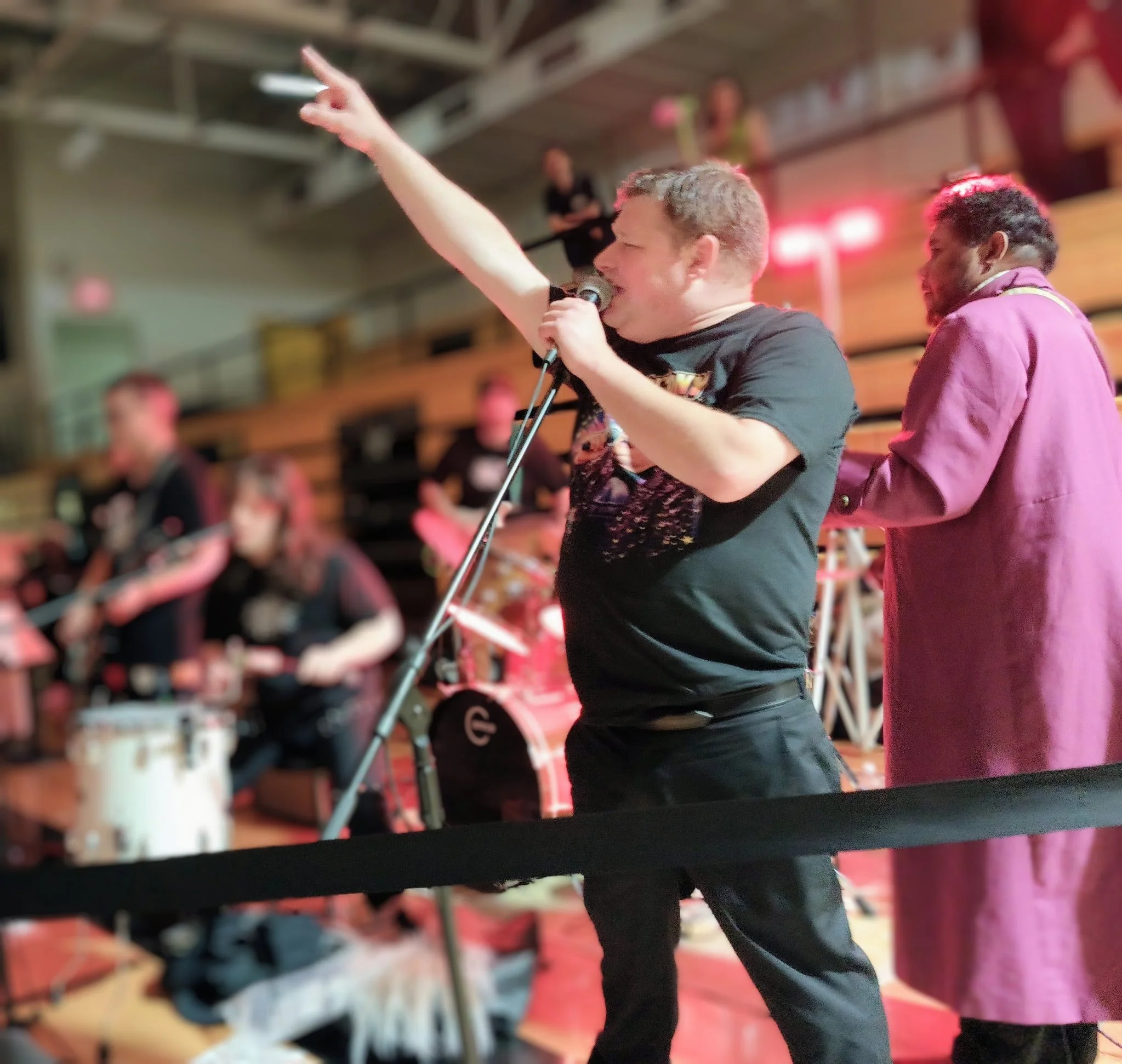 A person with short light hair and a black shirt singing into a microphone and raising their right hand in a lively indoor setting with blurred background and pink lighting.