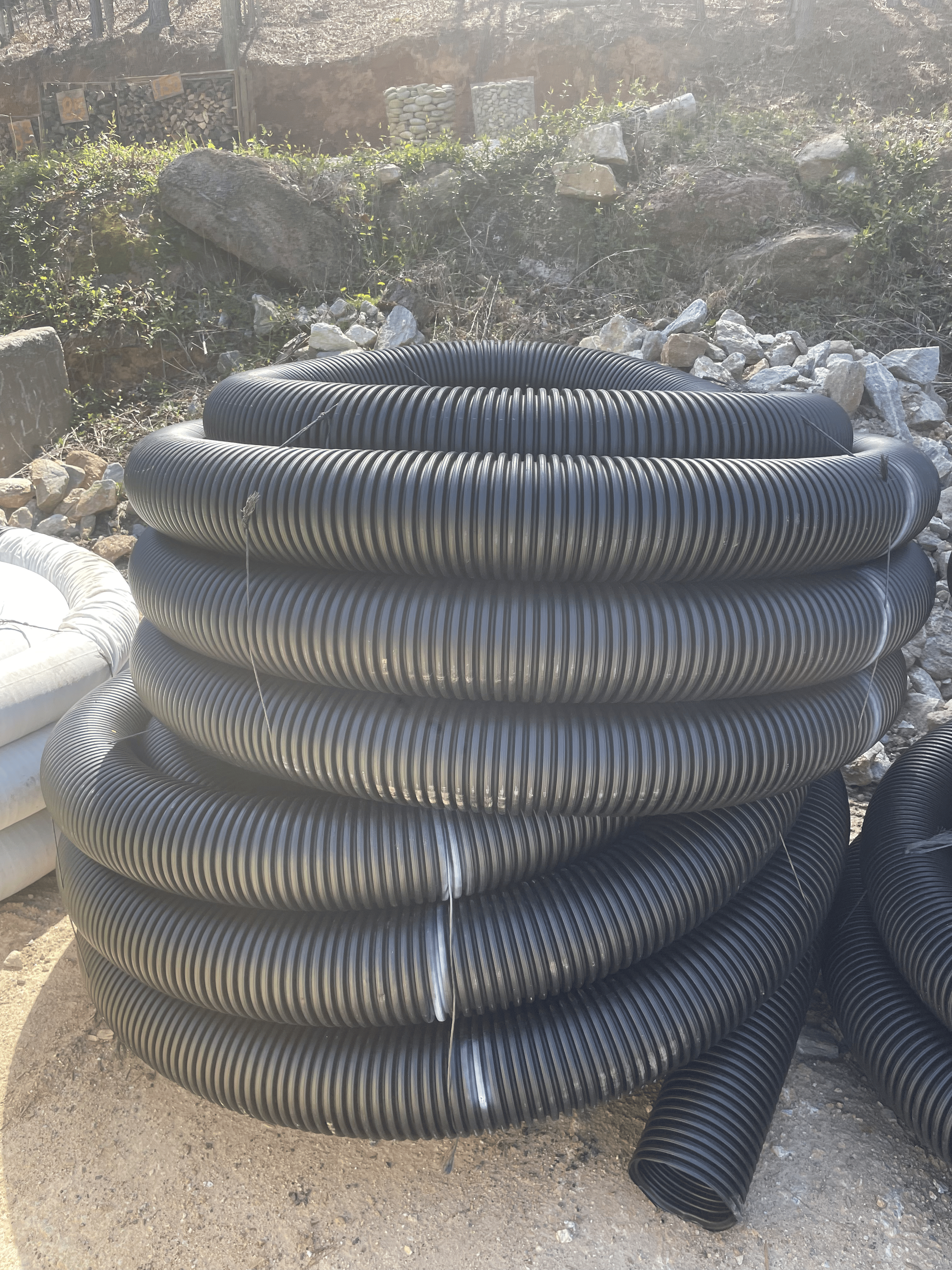 Stacked black corrugated pipes outdoors on dirt ground with rocks and unfinished landscape in the background.
