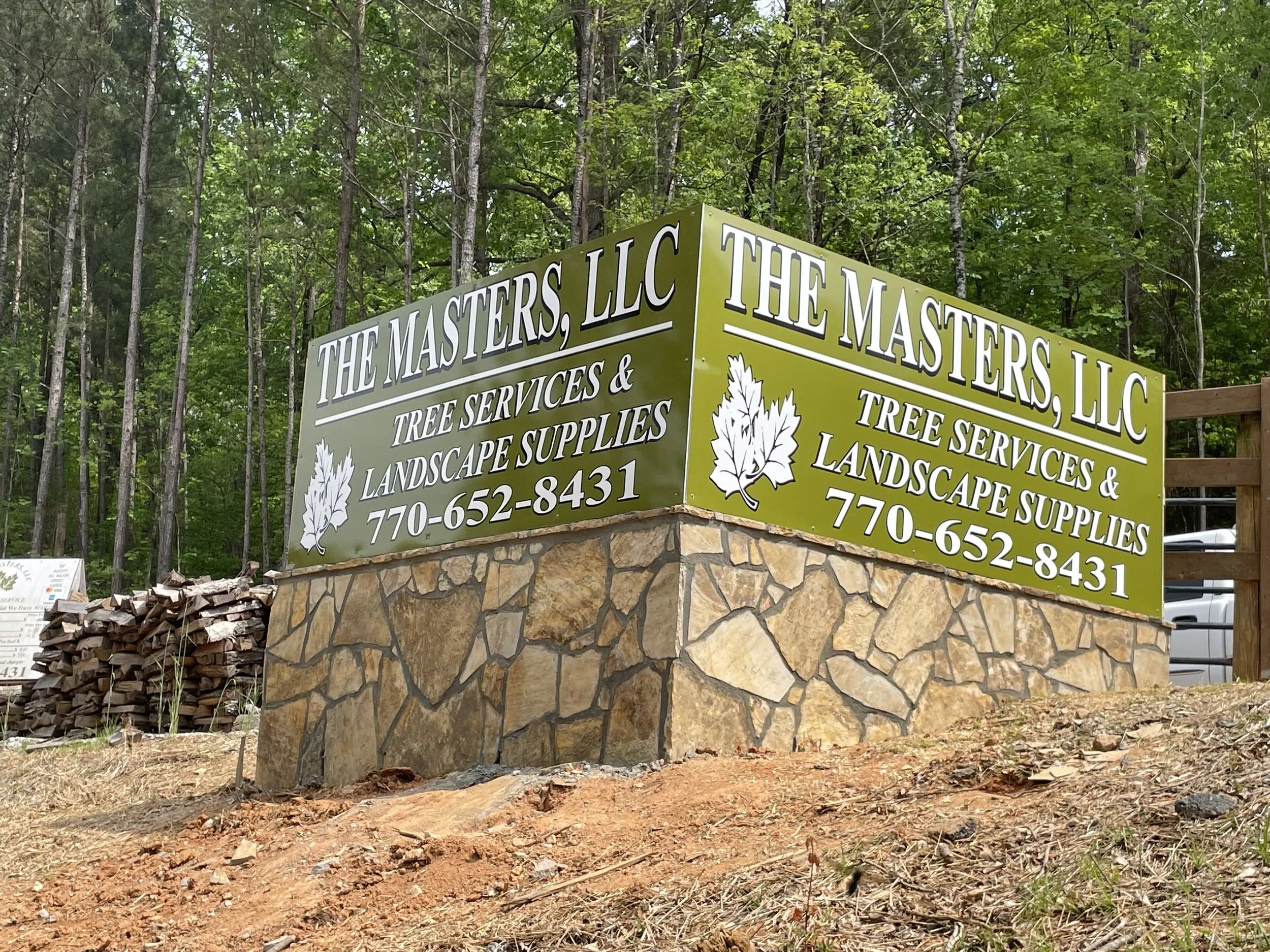 Sign for The Masters LLC, featuring services including tree services, landscape supplies, and landscaping, with a forest background and a stone foundation.