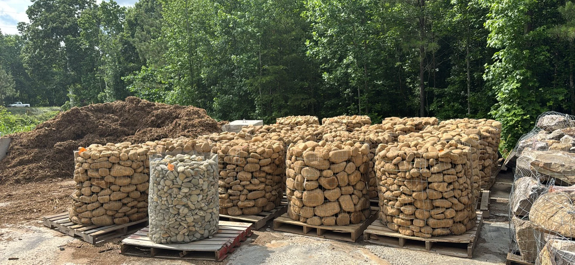 Stacks of large for landscaping that look like river rocks, stored on pallets outdoors with trees in the background.
