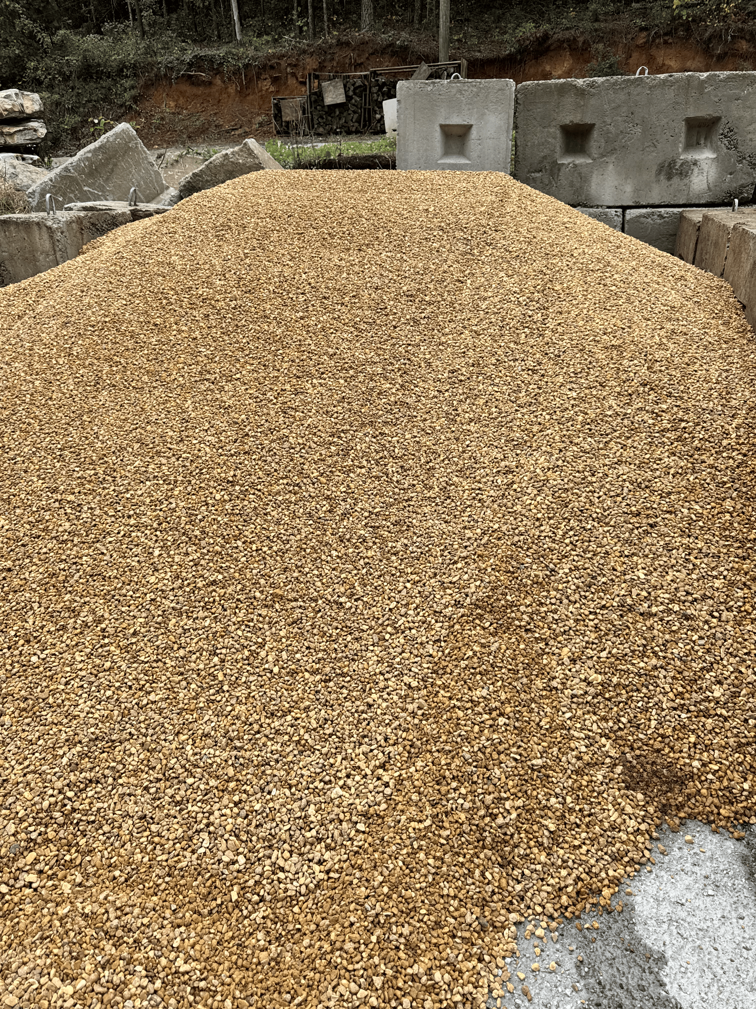 A large pile of small tan gravel at a construction site outdoors, with concrete blocks and a backdrop of trees and dirt.
