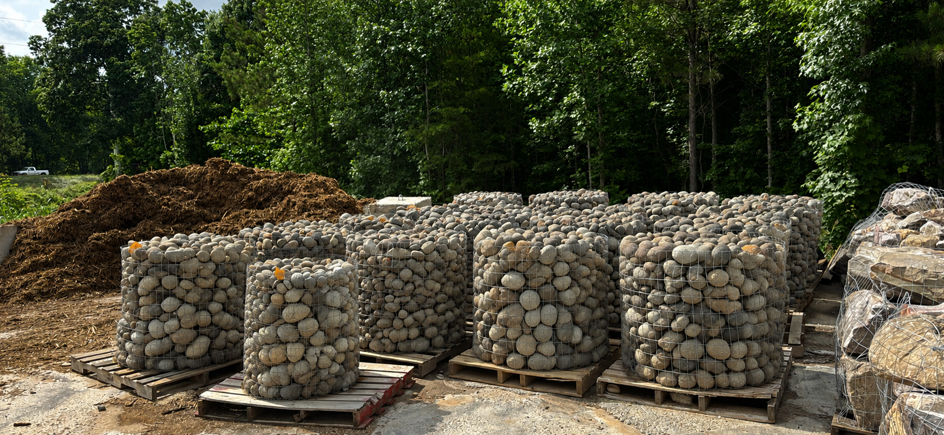 Multiple wire cages filled with assorted rocks placed on wooden pallets outdoors, with trees and a truck in the background.