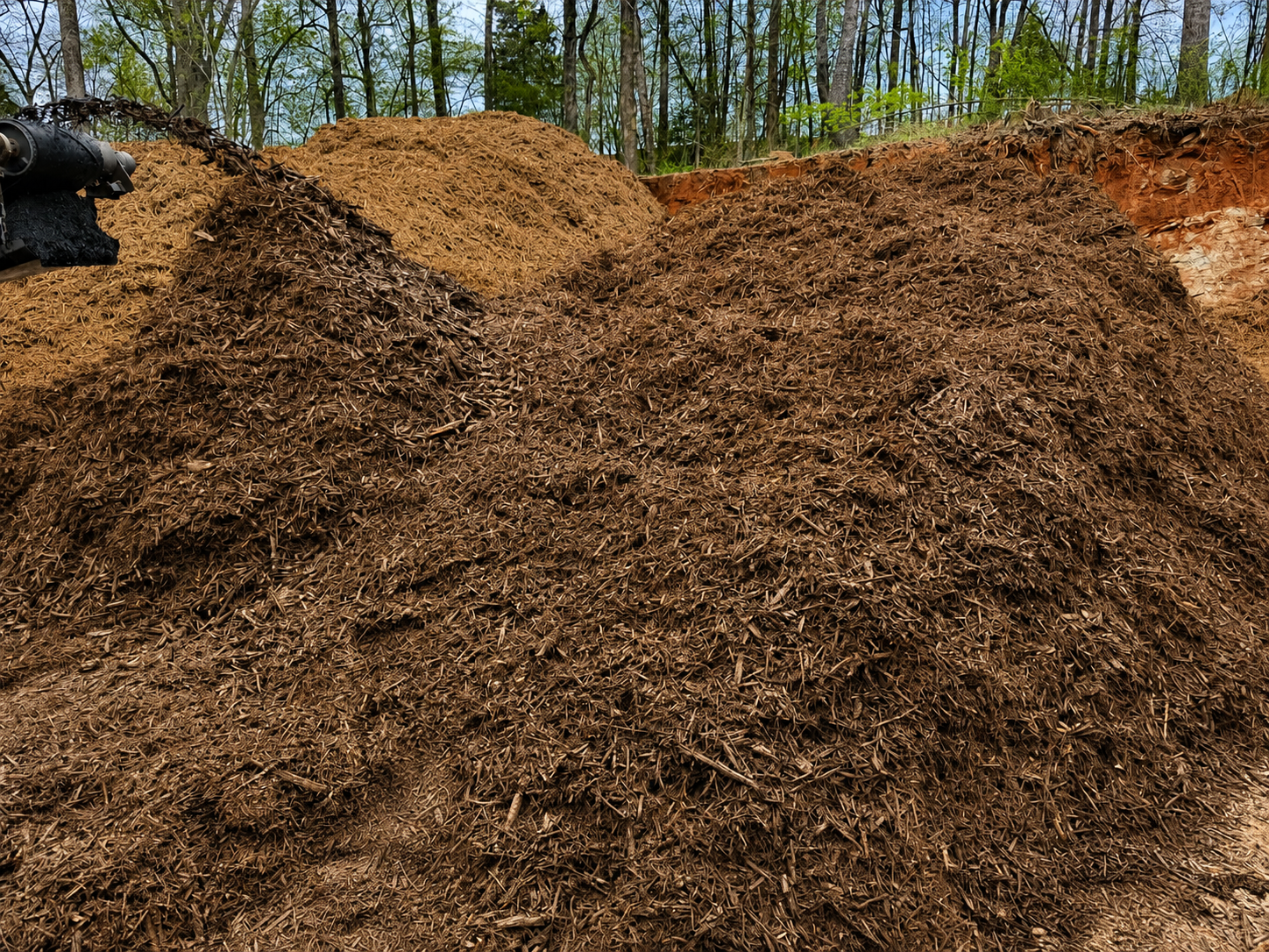 A pile of mulch in front of a wooded area with green trees and blue sky.