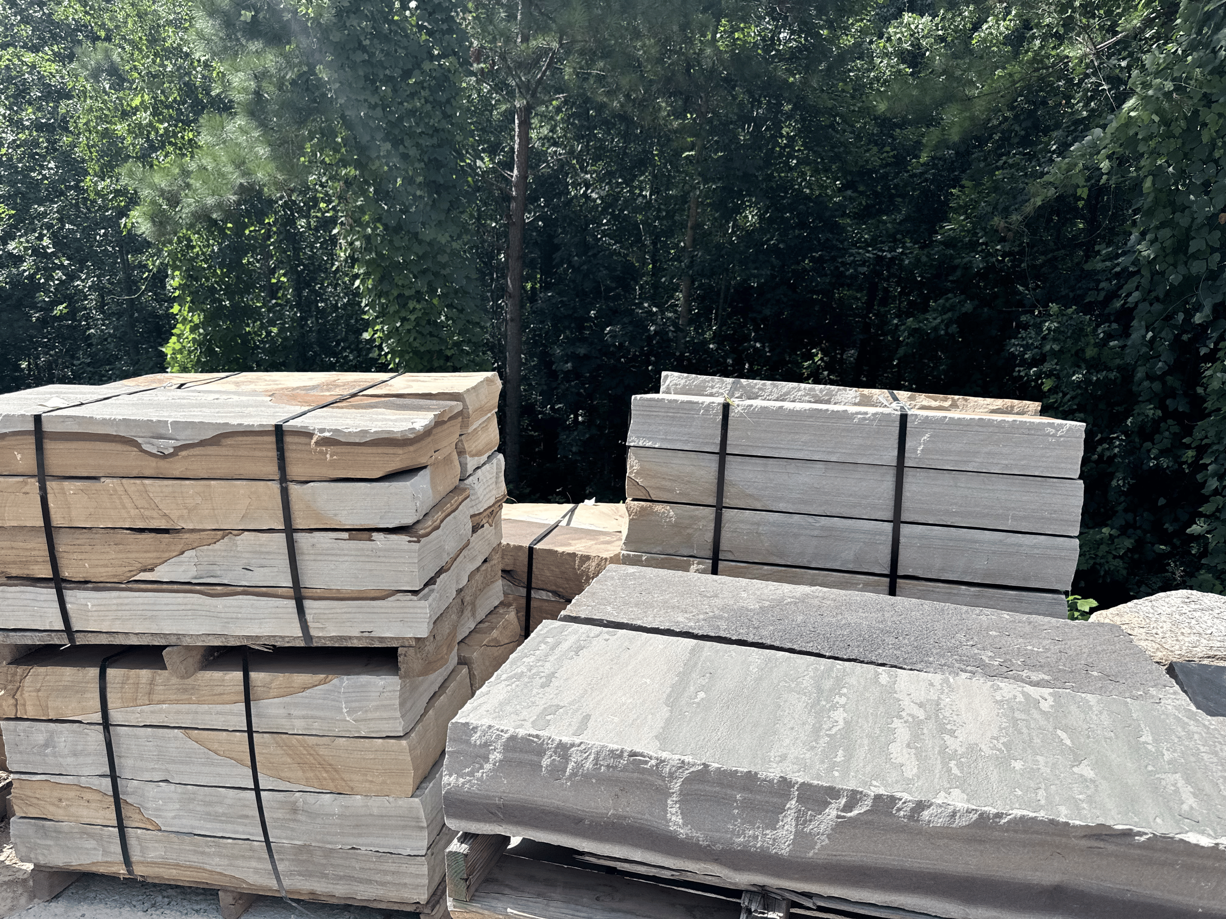 Stacks of flat stone tiles secured with black ties outdoors near dense trees.