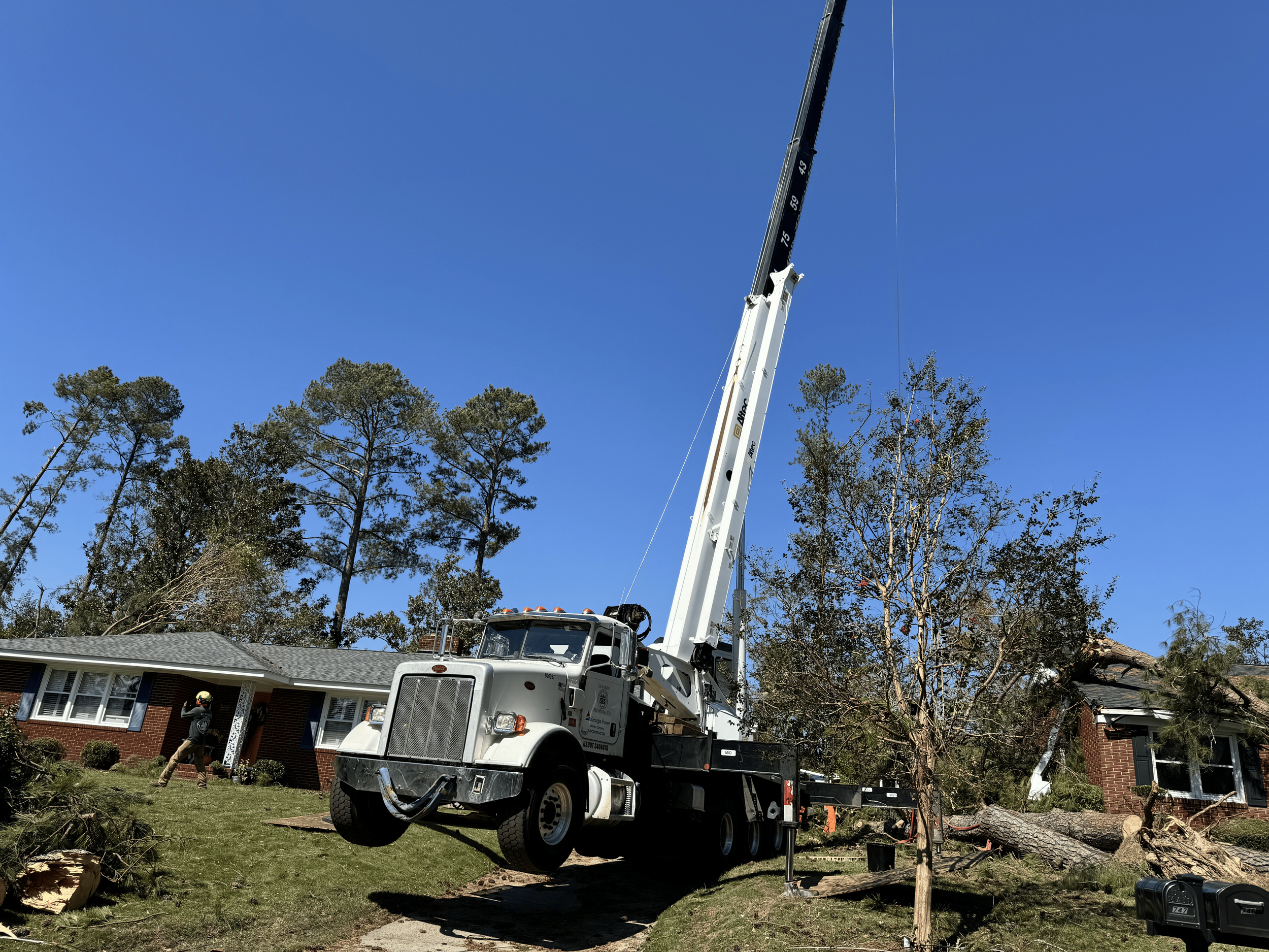 A large white crane truck is lifting a fallen tree onto a residential property, with two workers nearby managing the situation.