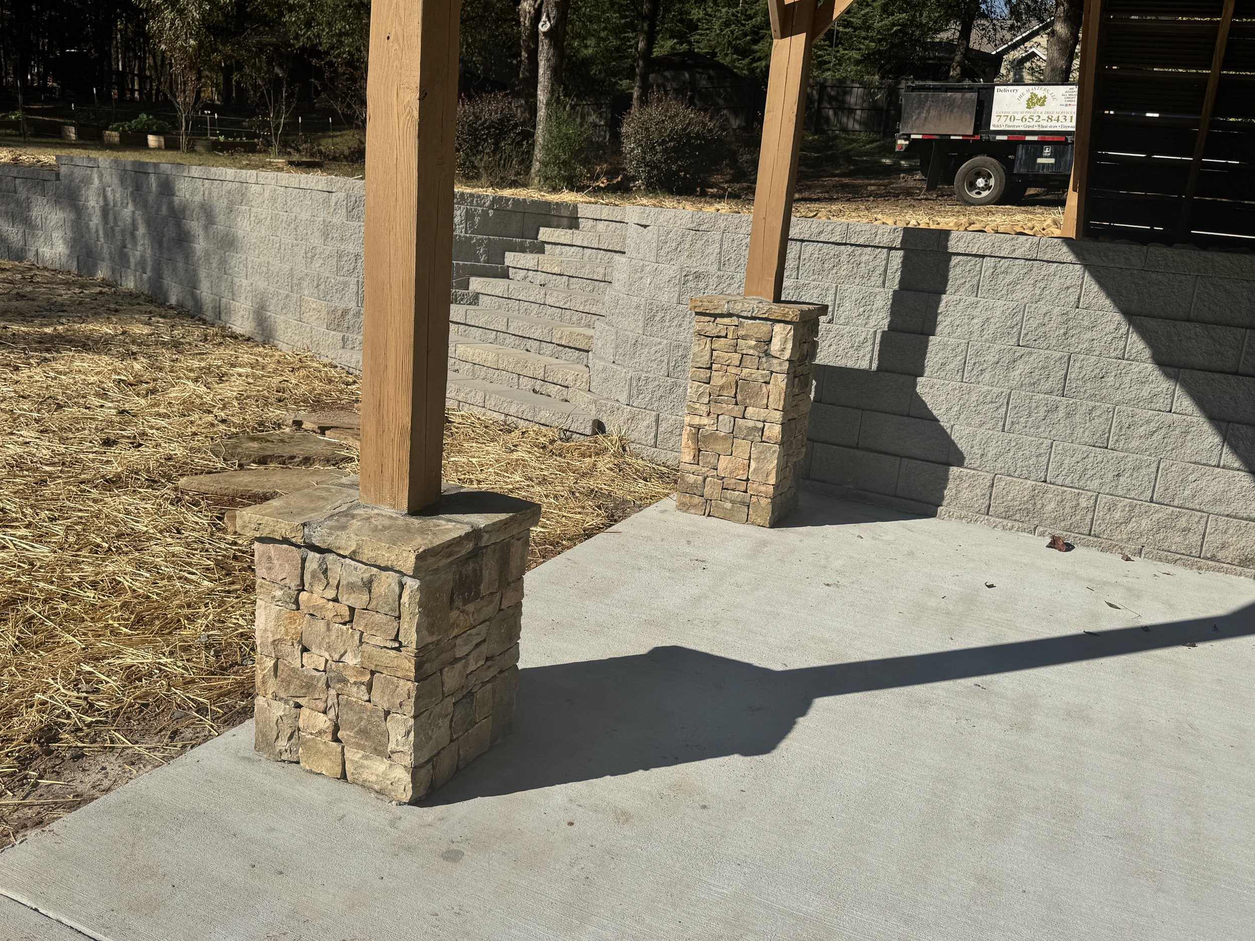 A newly constructed concrete patio with two stone pillar accents and orangewood posts supporting a structure. A stone retaining wall borders the space with a garden area beyond. A small black trailer with a sign is parked in the background, and trees and bushes are visible further back in a sunny outdoor setting.