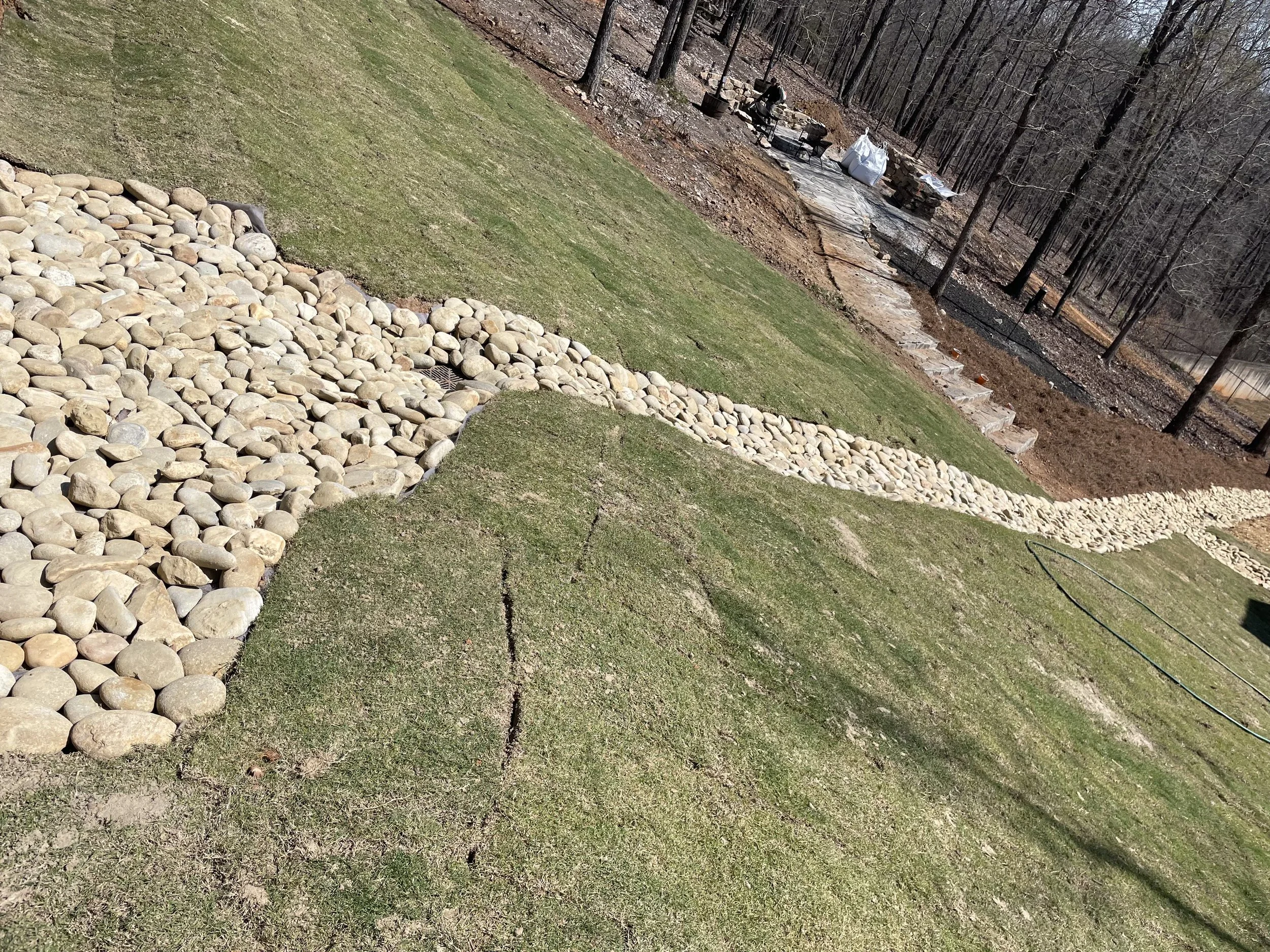 A backyard with a green lawn, a stone area, and a dirt path with some stones arranged in a border. In the background, there are trees with no leaves and a person sitting near a fire pit with supplies and furniture around.