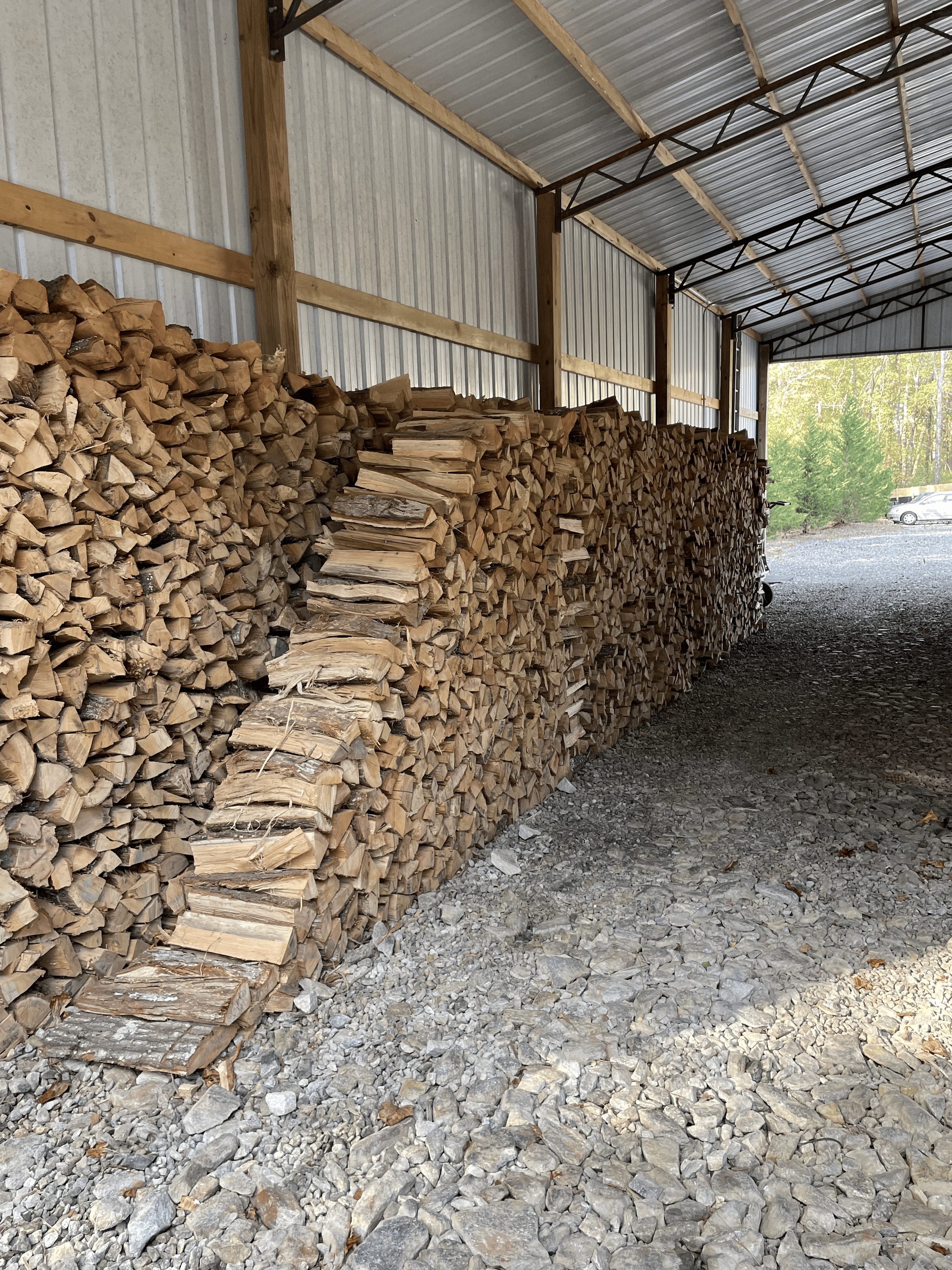Stacks of split firewood inside a shed with metal siding and roof, with a gravel ground and trees visible outside.