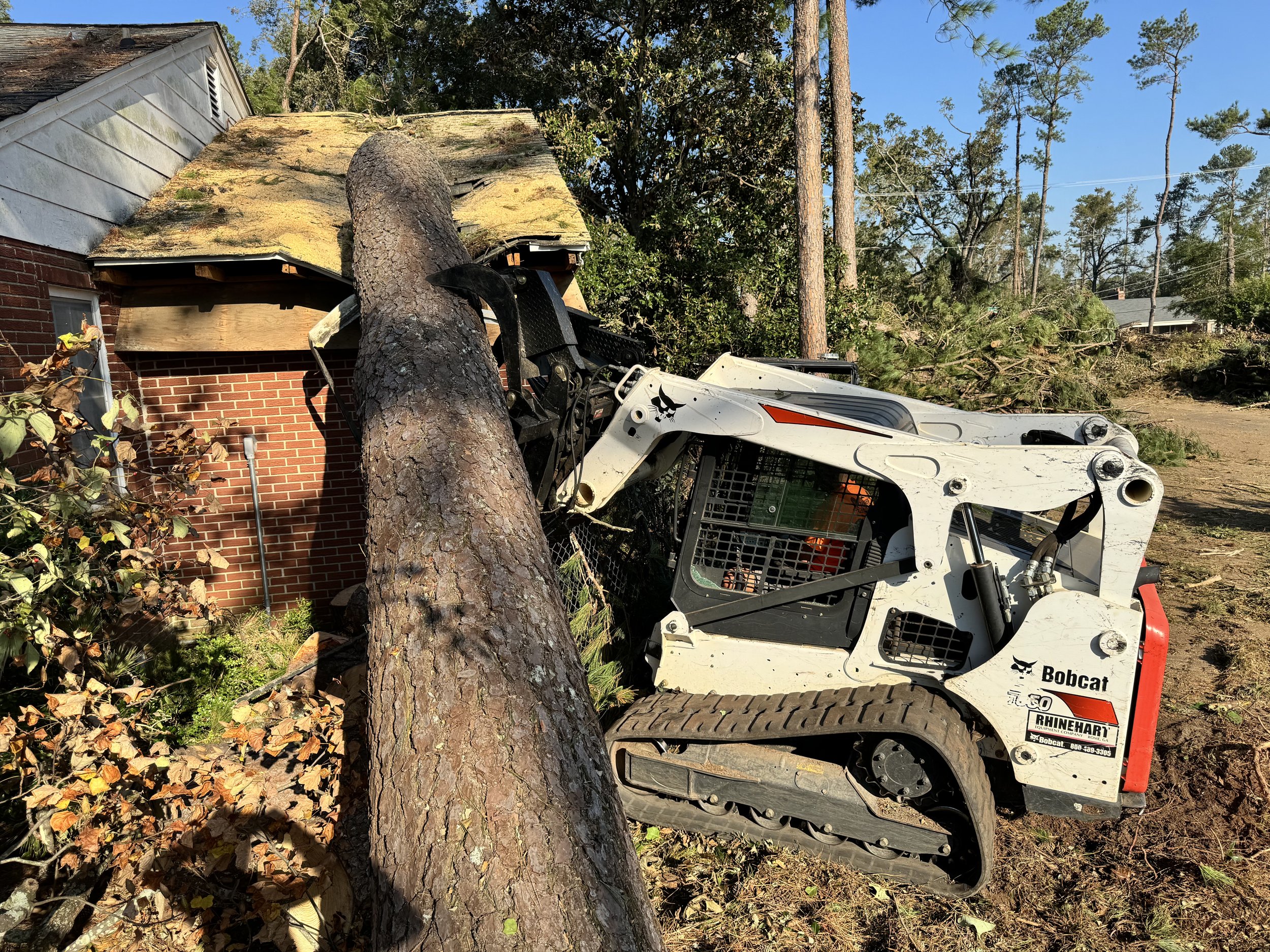 A large tree has fallen on a house, damaging the roof and exterior wall. A Bobcat construction vehicle is nearby, and debris from the tree and damage is scattered around.