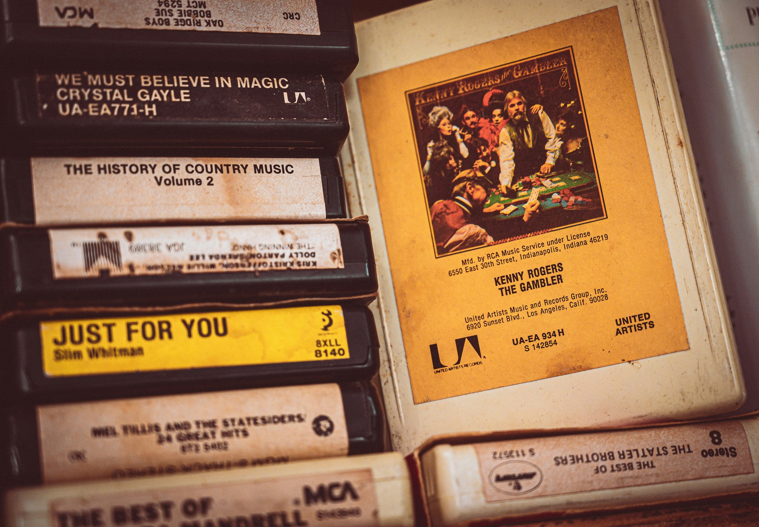 Close-up photograph of vintage cassette tapes stacked on a shelf, with some showing titles and labels. Notably, one cassette has a label titled 'WE MUST BELIEVE IN MAGIC' and 'CRYSTAL GAYLE,' and another titled 'THE HISTORY OF COUNTRY MUSIC Volume 2.' An orange cover of a record or album titled 'KENNY ROGERS THE GAMBLER' featuring an illustrated scene of people playing cards is also visible.