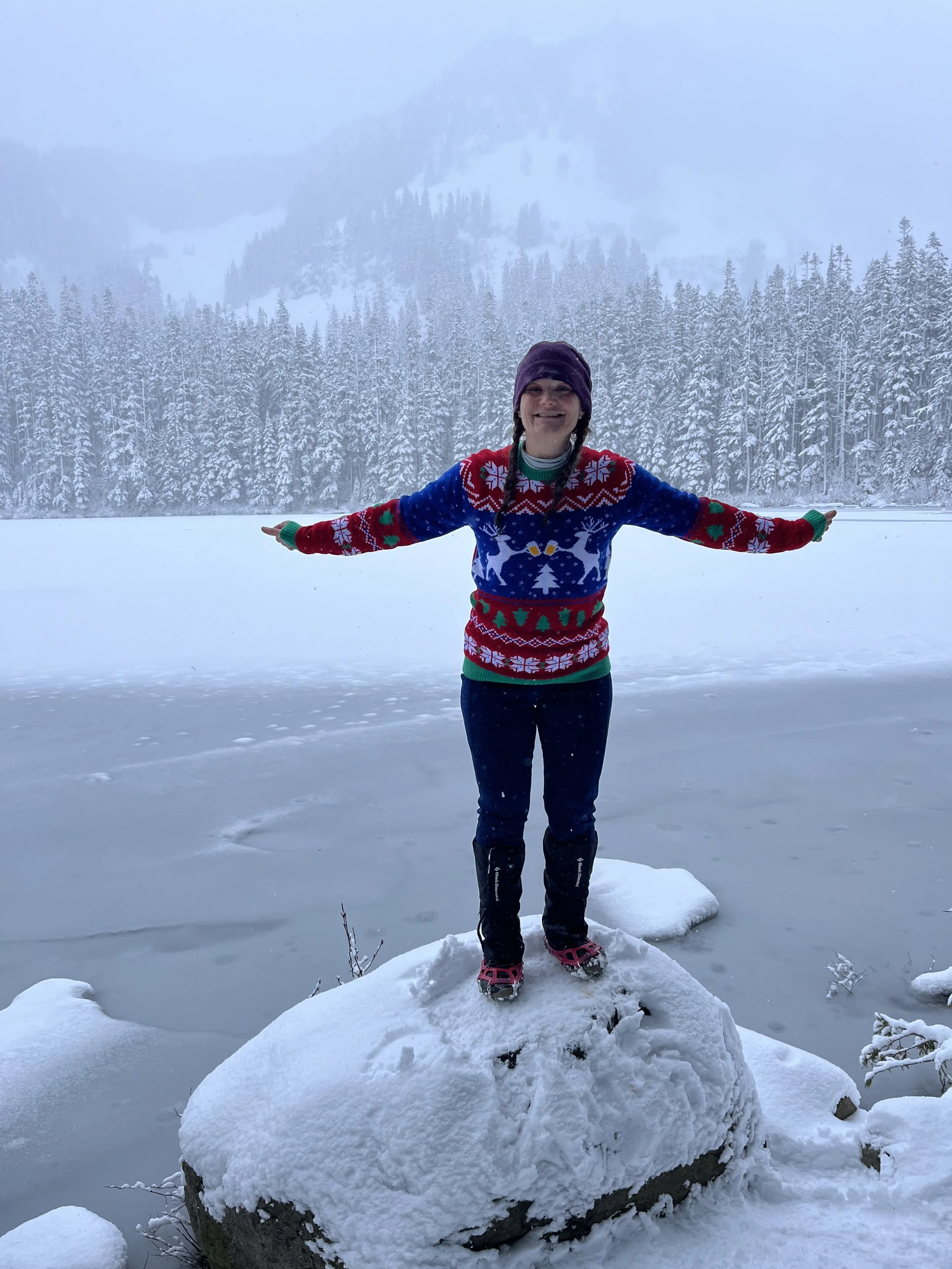 A person in winter clothing, including a colorful Christmas sweater, standing on a large snow-covered rock by a frozen lake with snow-covered trees and mountains in the background.