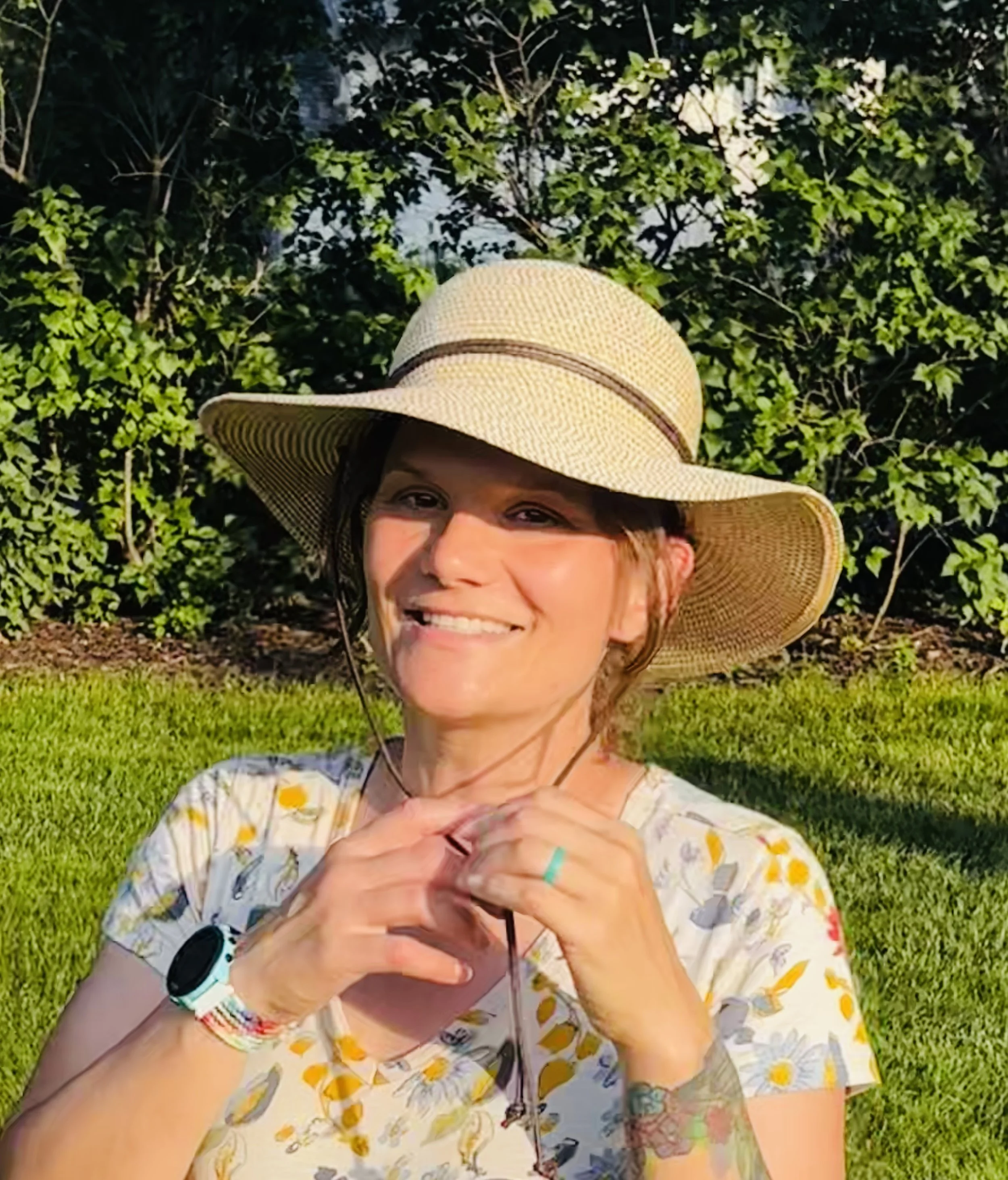 A woman wearing a straw wide-brimmed hat, sunglasses, and a floral shirt, smiling outdoors in front of green leafy bushes and grass on a sunny day.