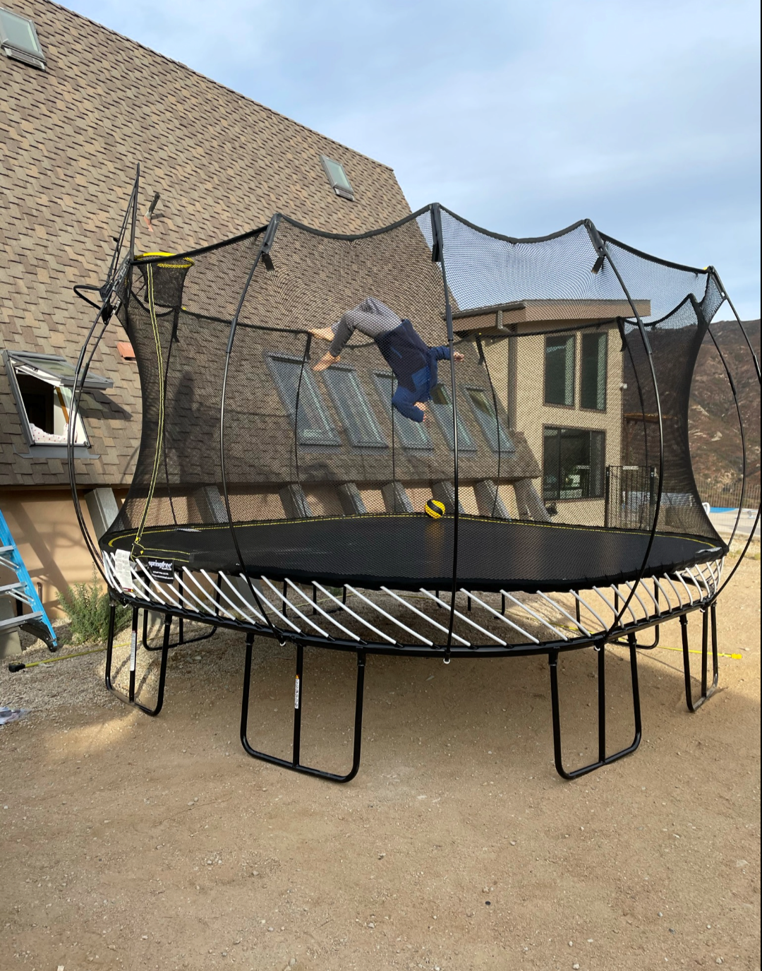    Person flipping on a large trampoline with a safety net near a modern house.