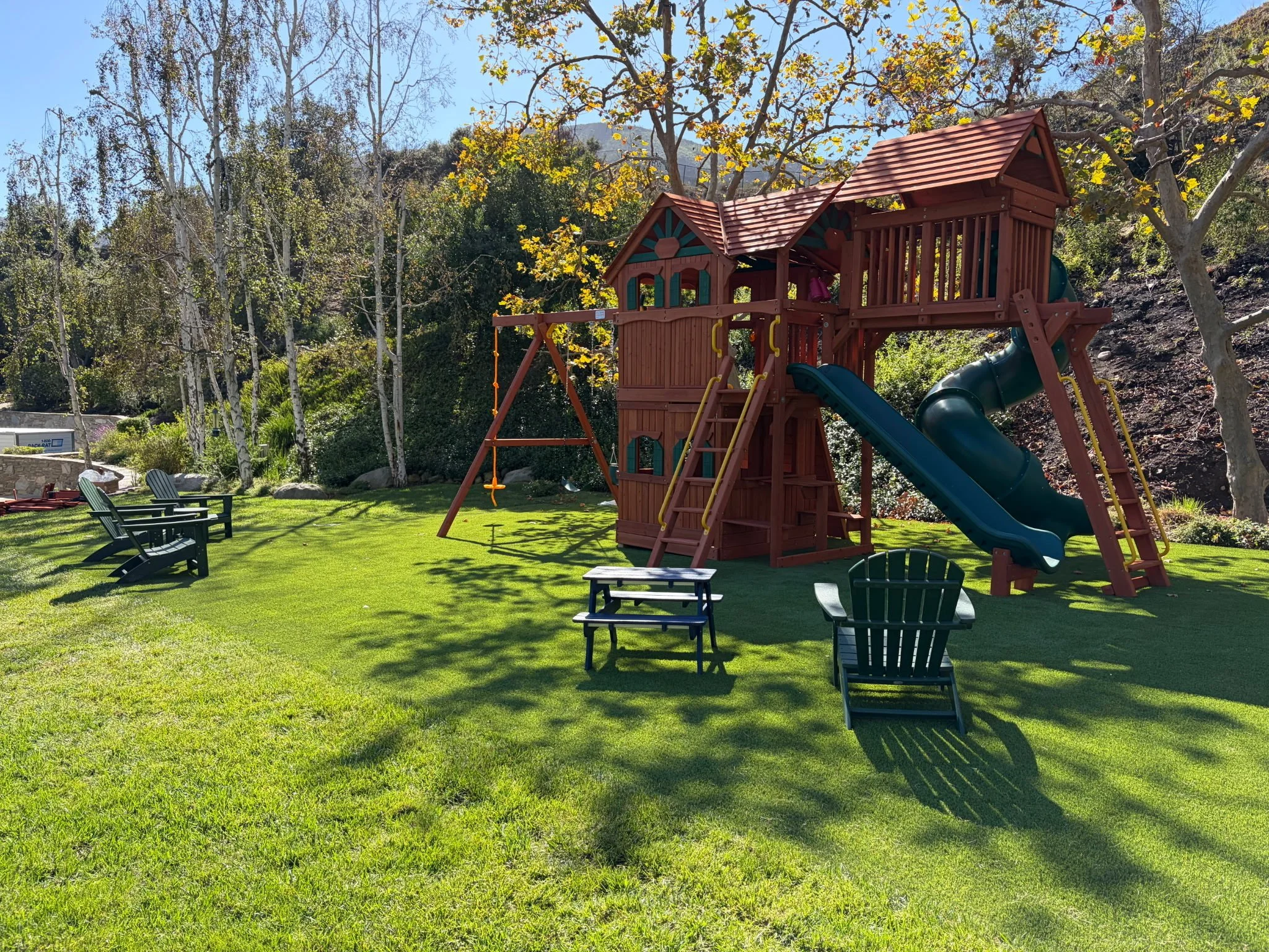 A wooden playground structure with a slide, swings, and climbing elements, situated on a grassy lawn surrounded by trees and outdoor seating.