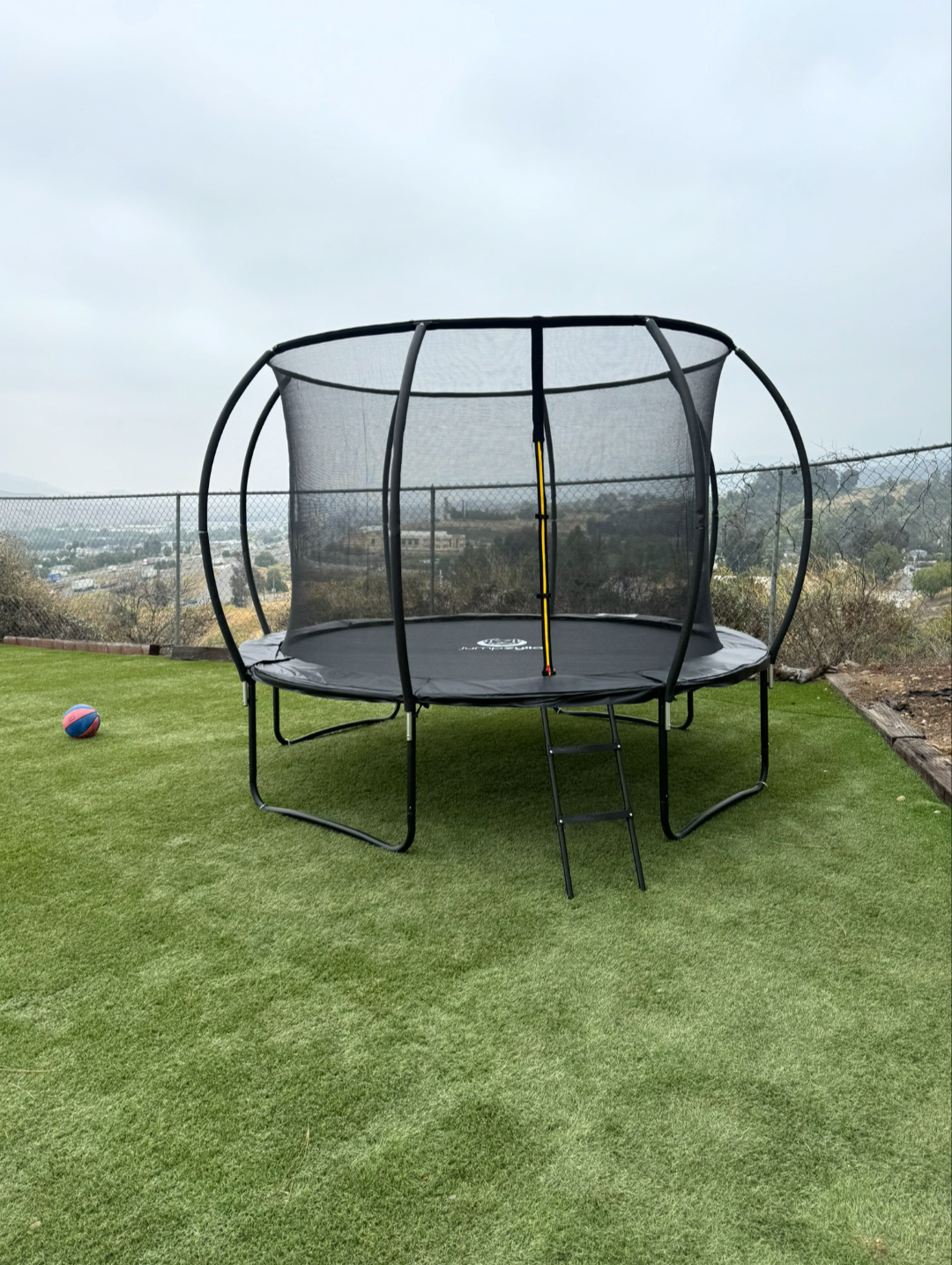    Round trampoline with a safety net on artificial grass near a chain‑link fence.