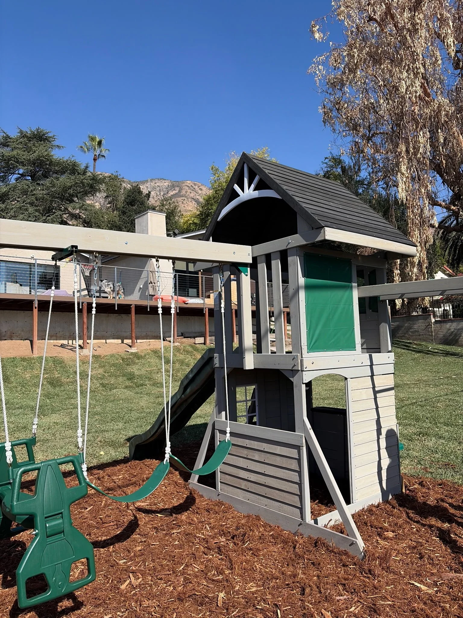 Backyard playset with a small tower, slide, and swings on mulch.