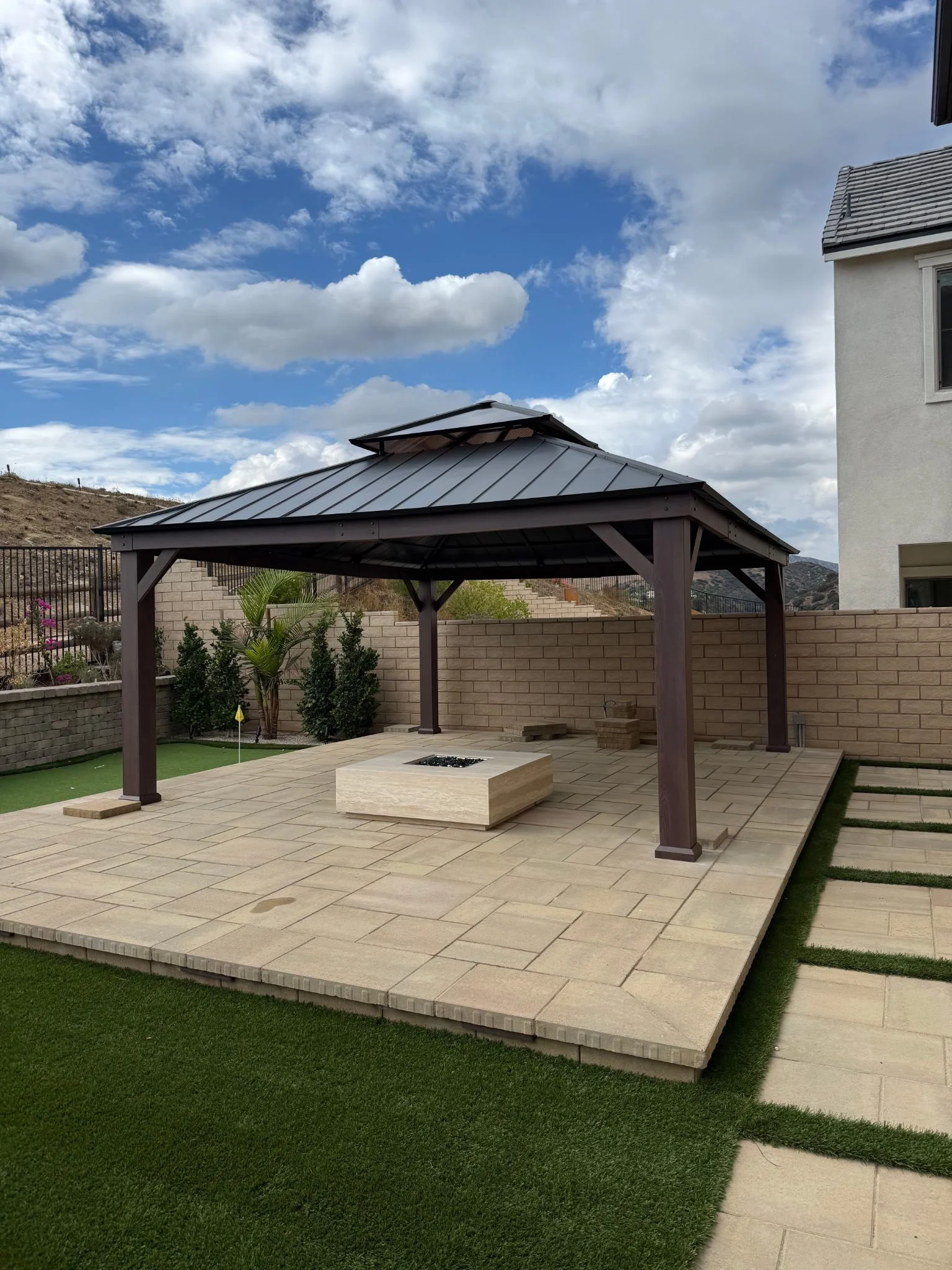 Backyard patio with a wooden gazebo, fire pit, stone flooring, and artificial grass.