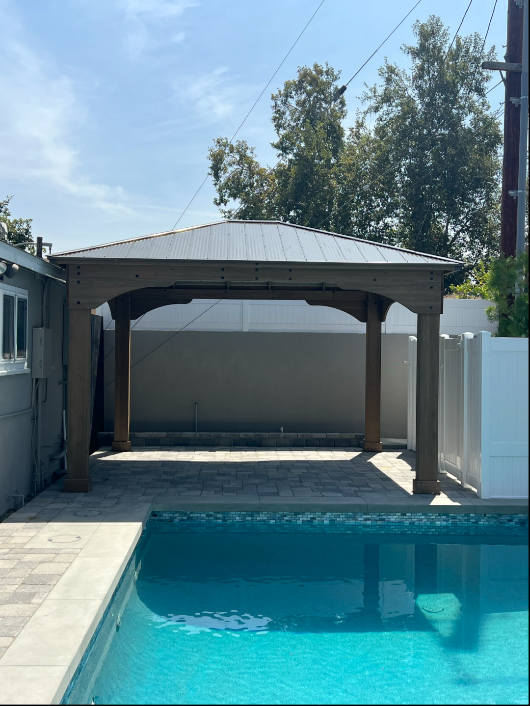 Poolside gazebo with a pitched roof on a paved patio beside a house.