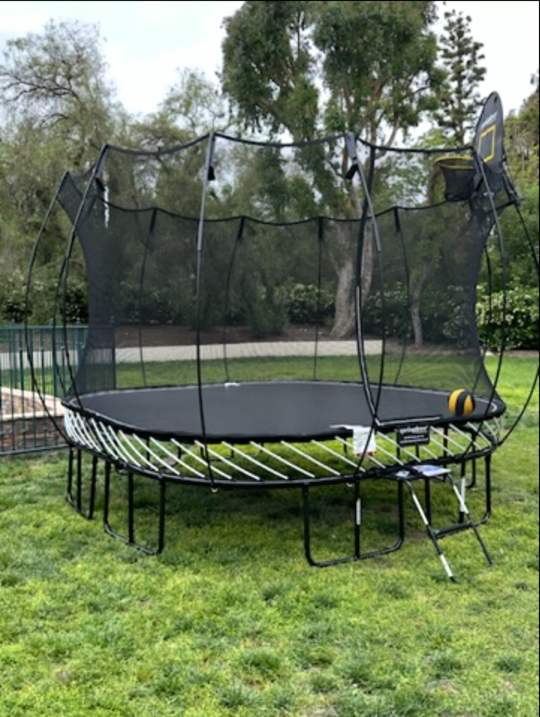 Round trampoline with a safety net on natural grass surrounded by trees.
