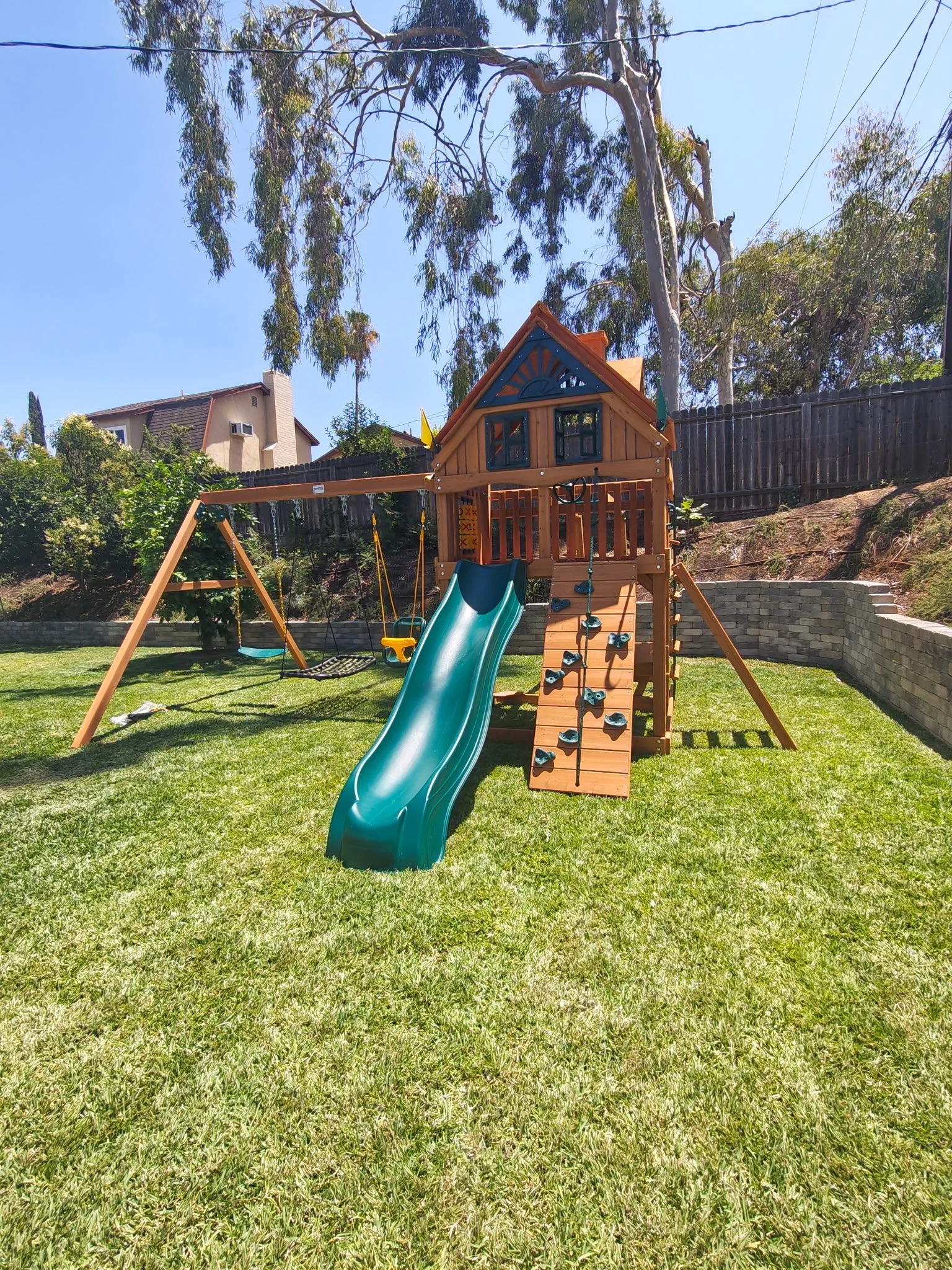 Backyard playset with a slide, climbing wall, swings, and a small roofed tower.