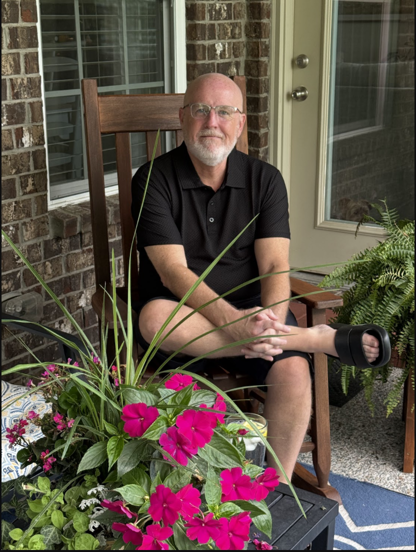 A man with glasses and a beard, wearing a black polo shirt, sitting outside on a wooden chair on a porch. He has one leg crossed over the other, with his right foot resting on his left knee, and his hands clasped. There are pink flowers and green plants in front of him, with a brick wall, window, and glass door in the background.