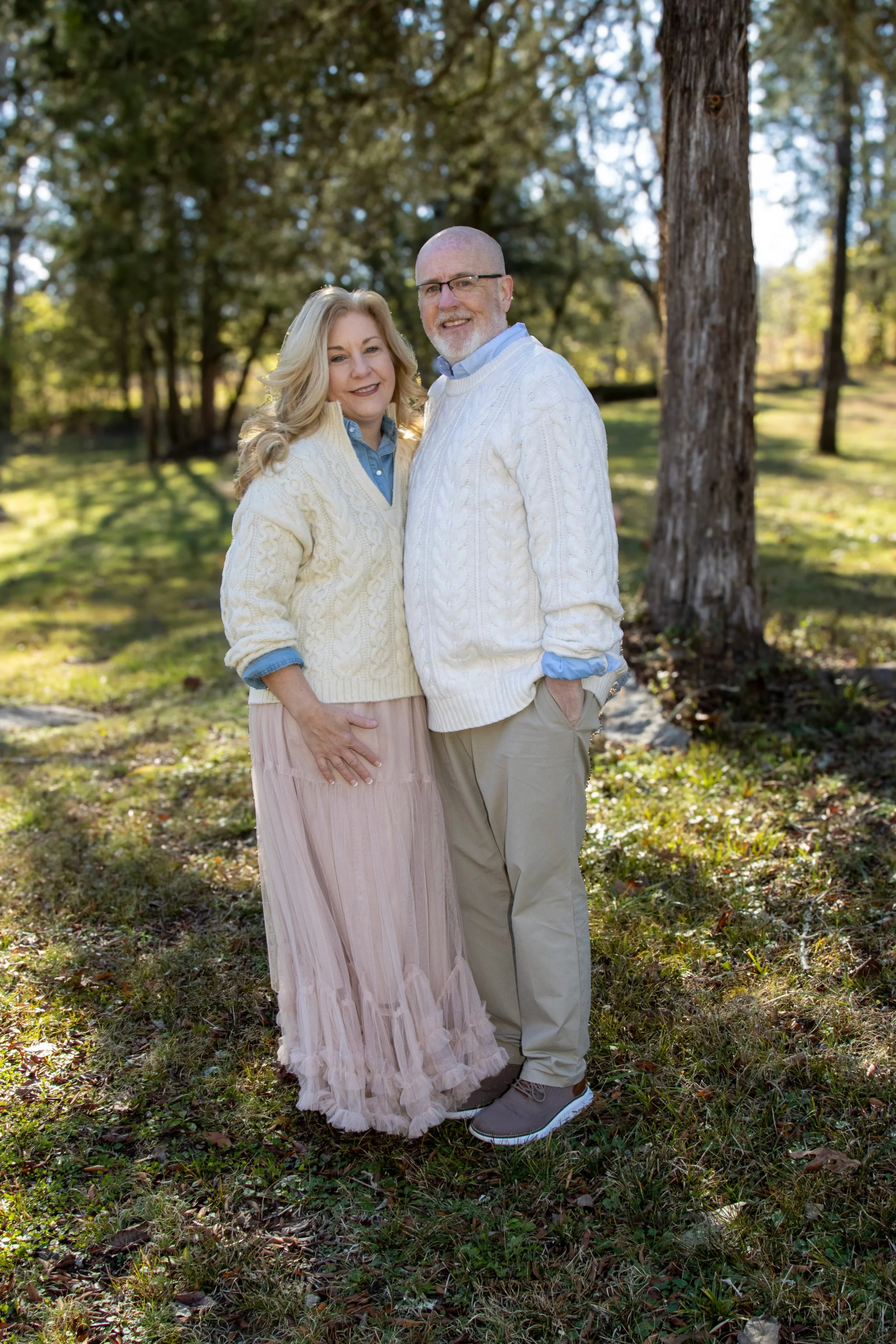 A middle-aged couple standing outdoors in a wooded area with autumn-colored trees, smiling at the camera. The woman has long blonde hair and is wearing a cream knit sweater over a blue shirt and a long pink pleated skirt. The man has a beard, glasses, and is wearing a cream cable-knit sweater over a blue collared shirt and khaki pants.