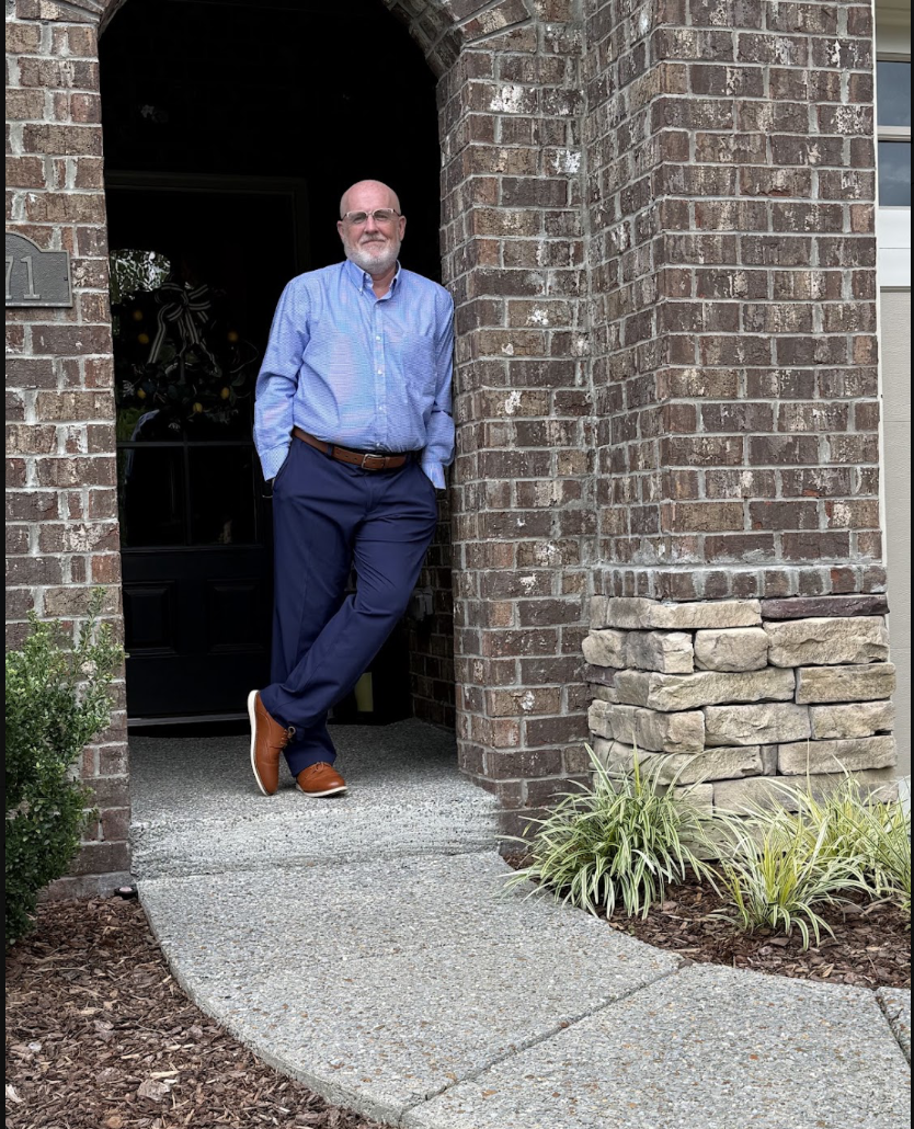 A man with glasses and a beard, dressed in a blue shirt and navy pants, standing casually at the entrance of a brick house.