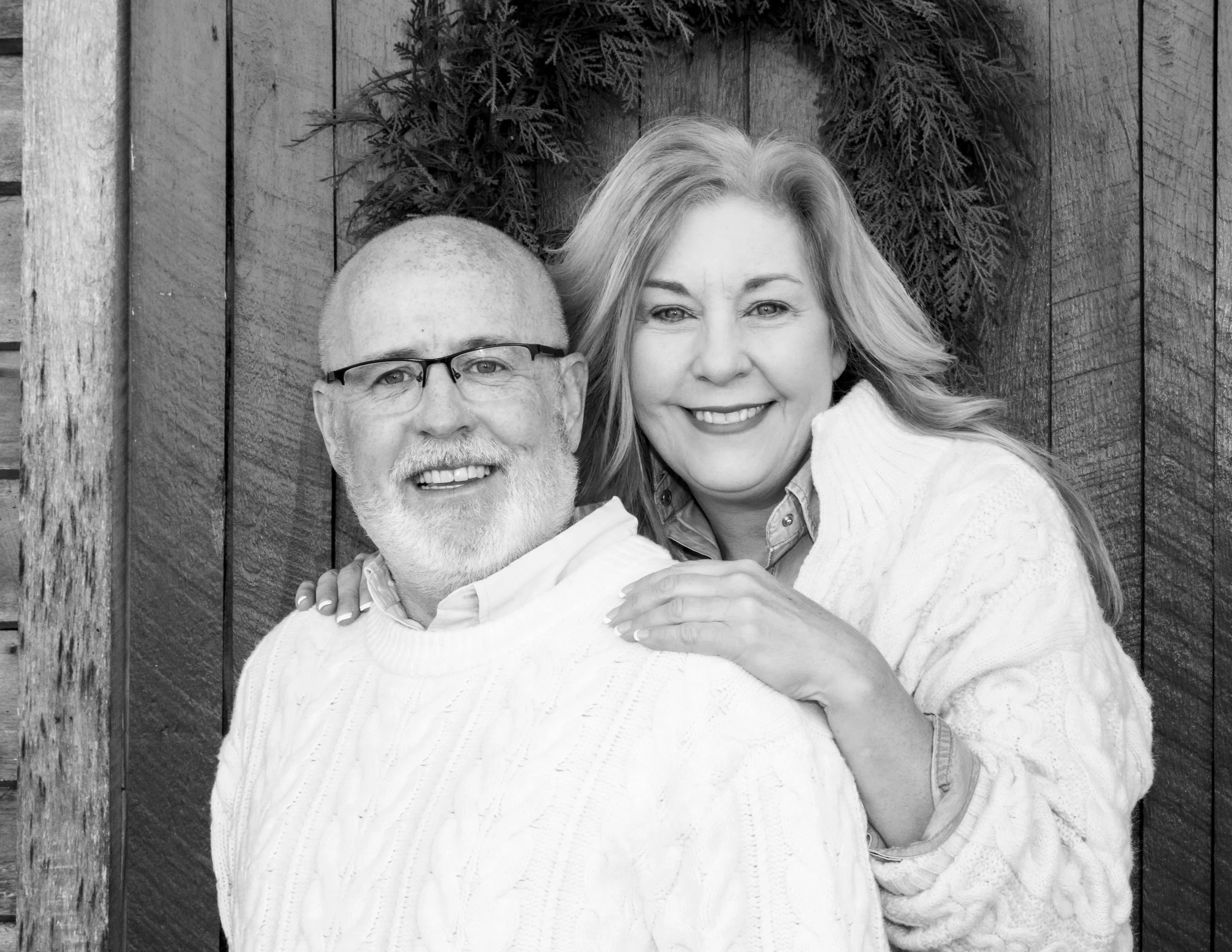 A smiling couple, an older man with glasses and a white beard, and a woman with blonde hair, in a close embrace in front of a wooden wall with a wreath and evergreen branches.
